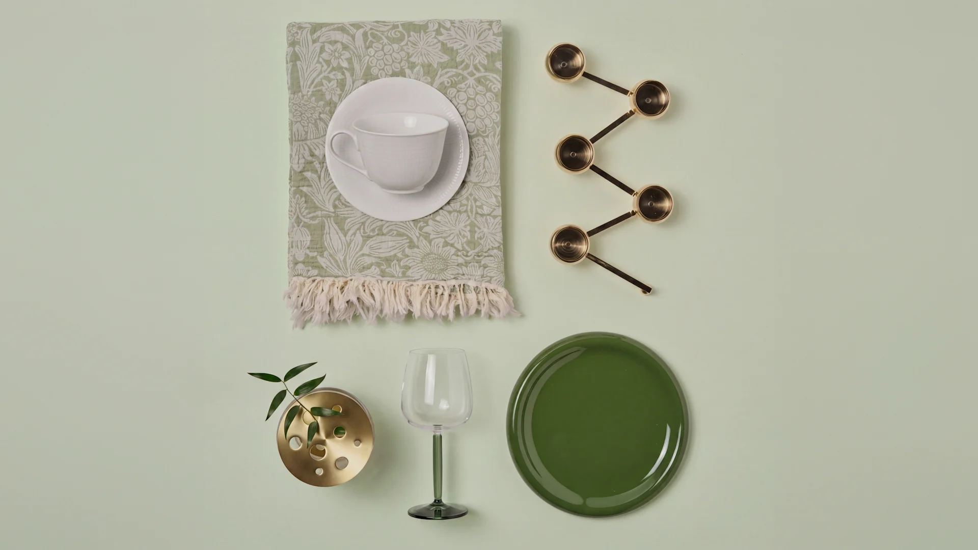 Tazza bianca, tovagliolo verde, misurini ottone, vaso pianta, calice vino verde, piatto verde scuro.