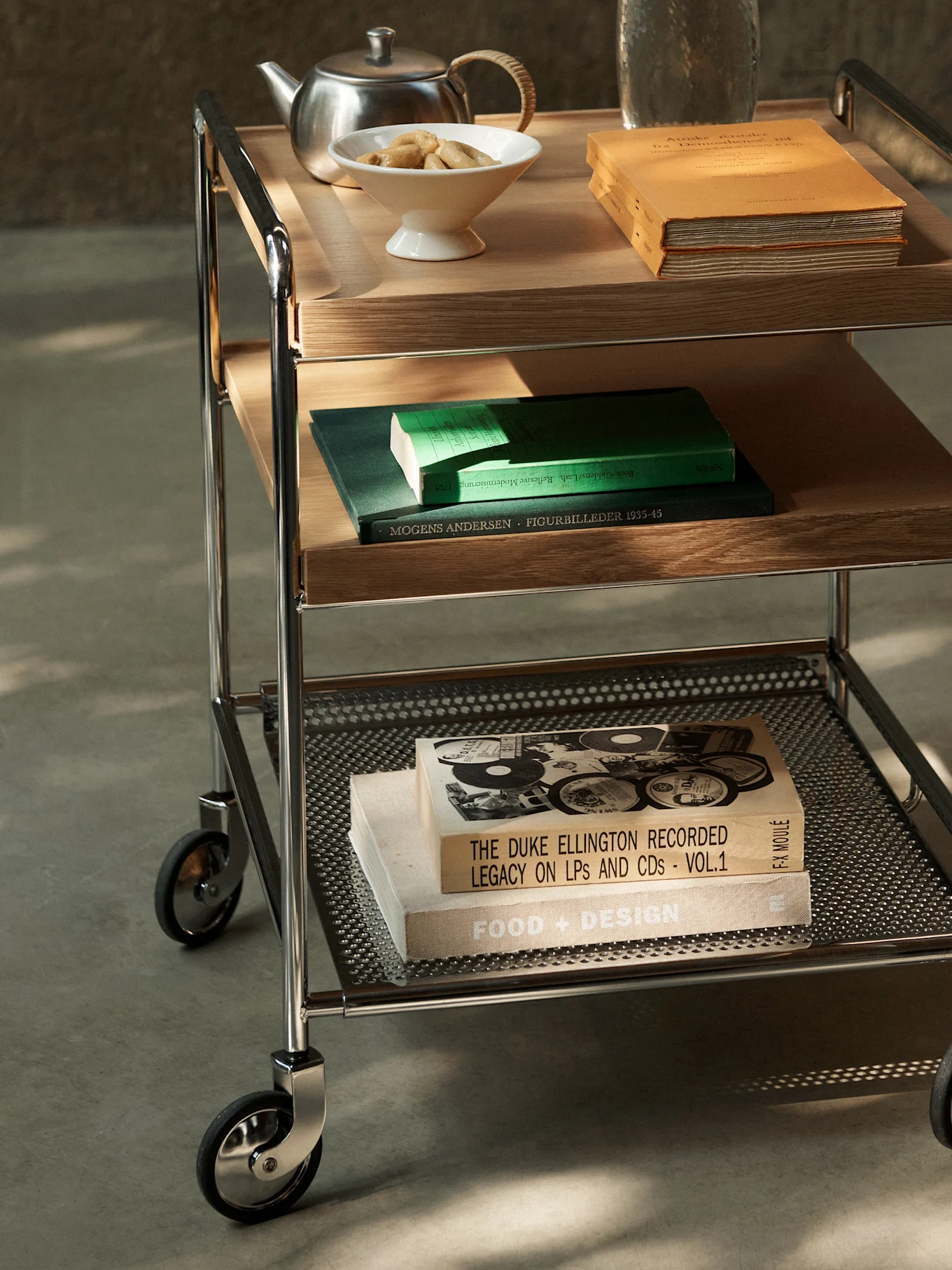 Un carrello di servizio a tre ripiani con struttura cromata, ripiani in legno e metallo perforato, contenente libri, teiera e biscotti.