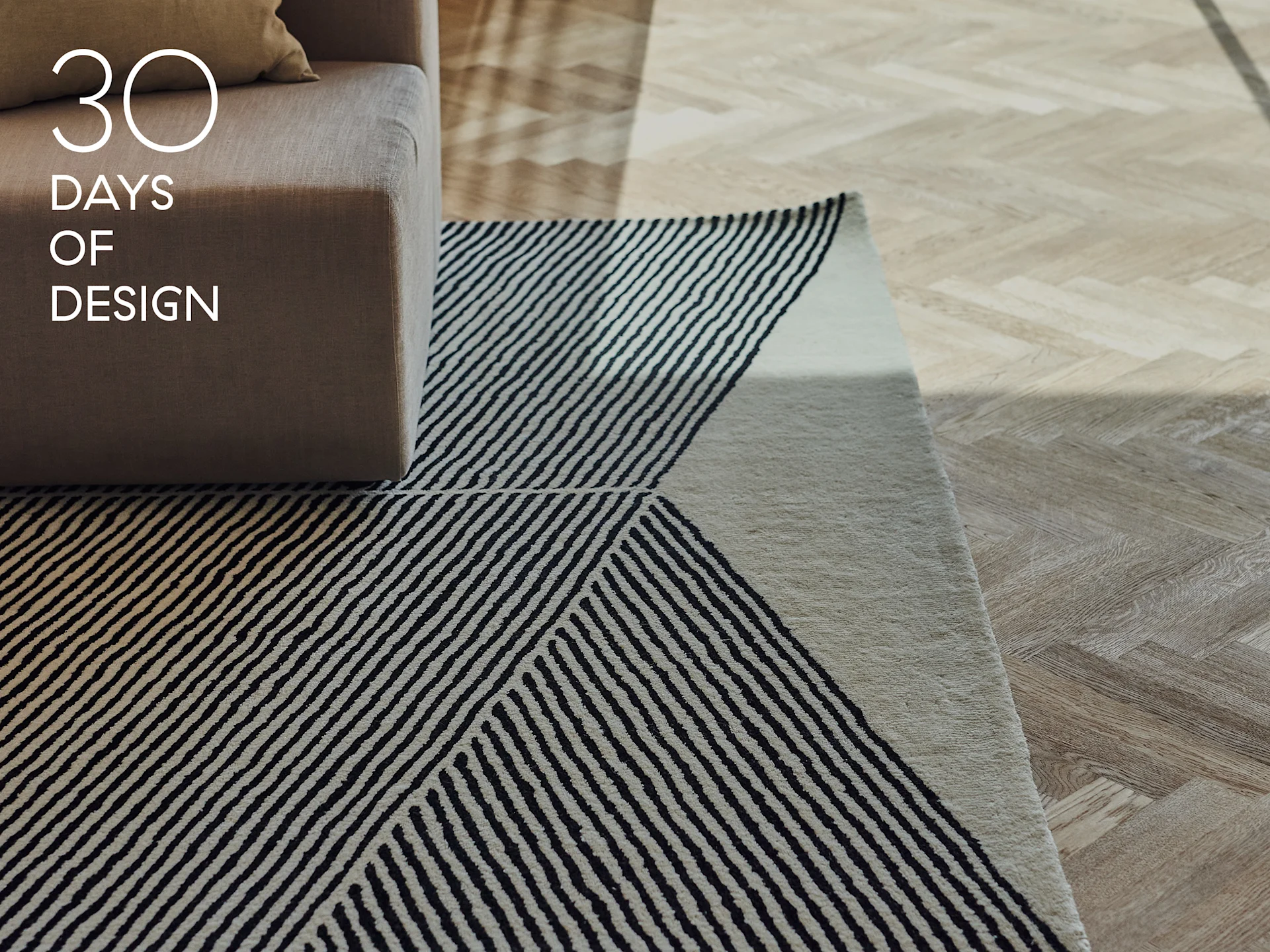 Bracciolo del divano, tappeto a strisce geometriche bianche e nere e pavimento in legno chiaro a spina di pesce. Testo: 30 DAYS OF DESIGN.