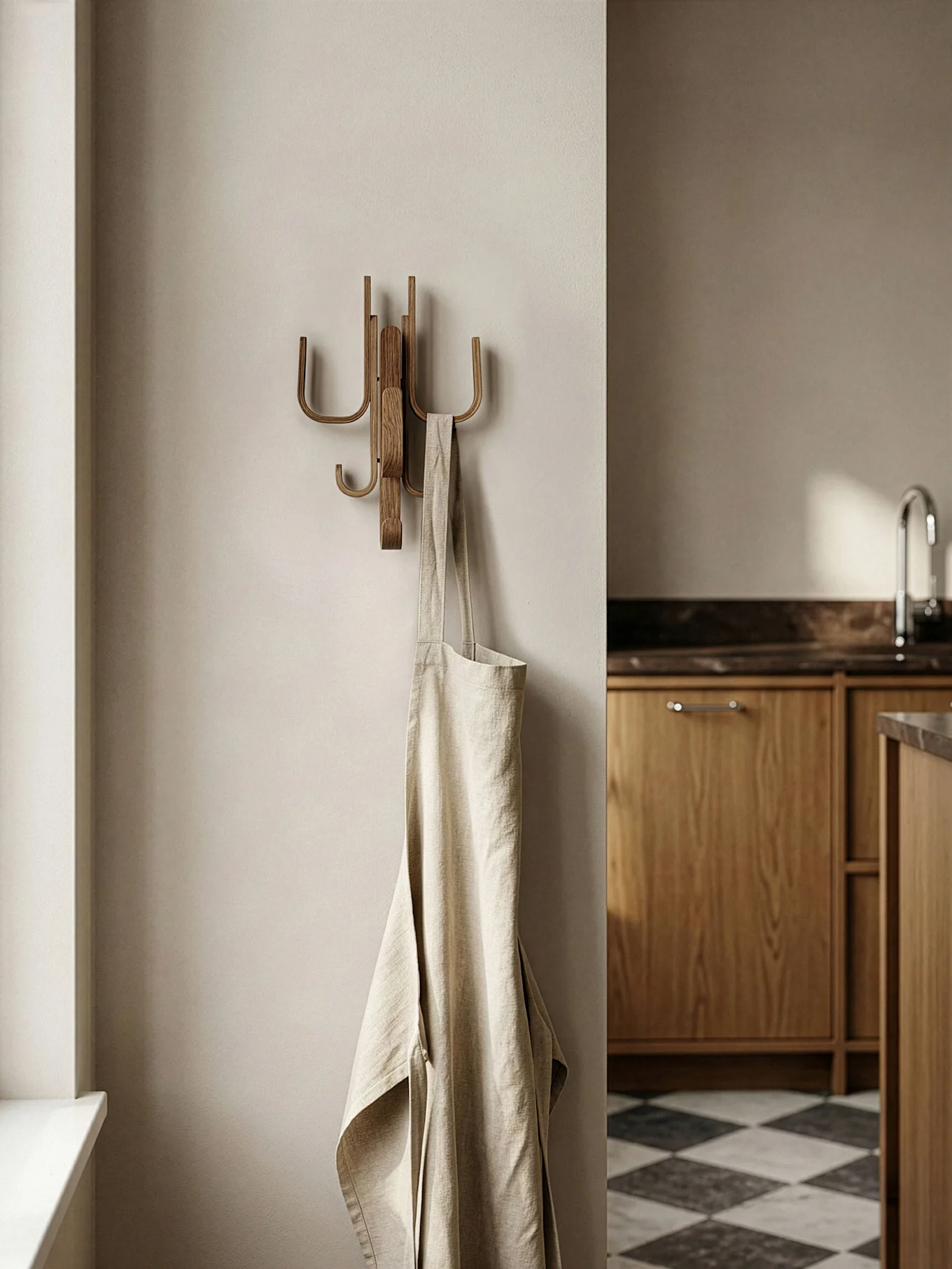 Un grembiule di lino beige chiaro pende da un appendiabiti in legno in una cucina moderna con mobili in legno e pavimento a scacchi.