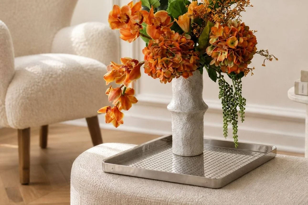 Vivaci fiori arancioni in un vaso bianco testurizzato su un vassoio d'argento, posizionati su un pouf chiaro in un soggiorno moderno.