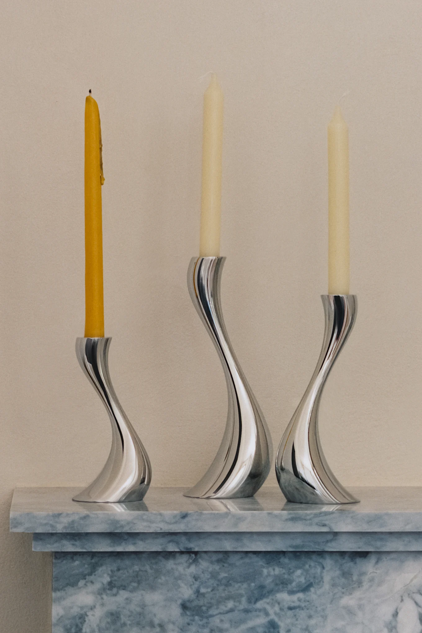 Tre eleganti portacandele argentati e curvi con una candela gialla bruciata e due candele color crema non accese su una superficie di marmo.