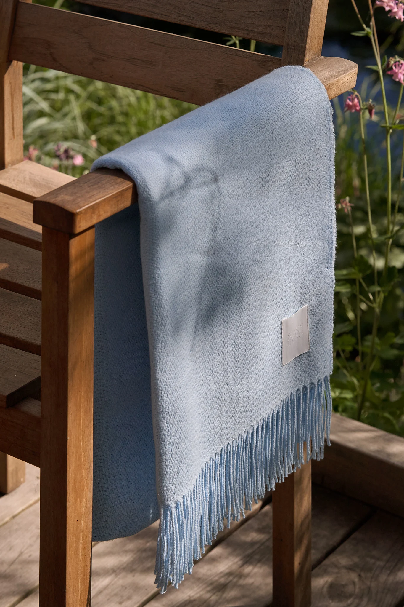 Coperta azzurra con frange drappeggiata su una sedia da esterno in legno in un giardino soleggiato.