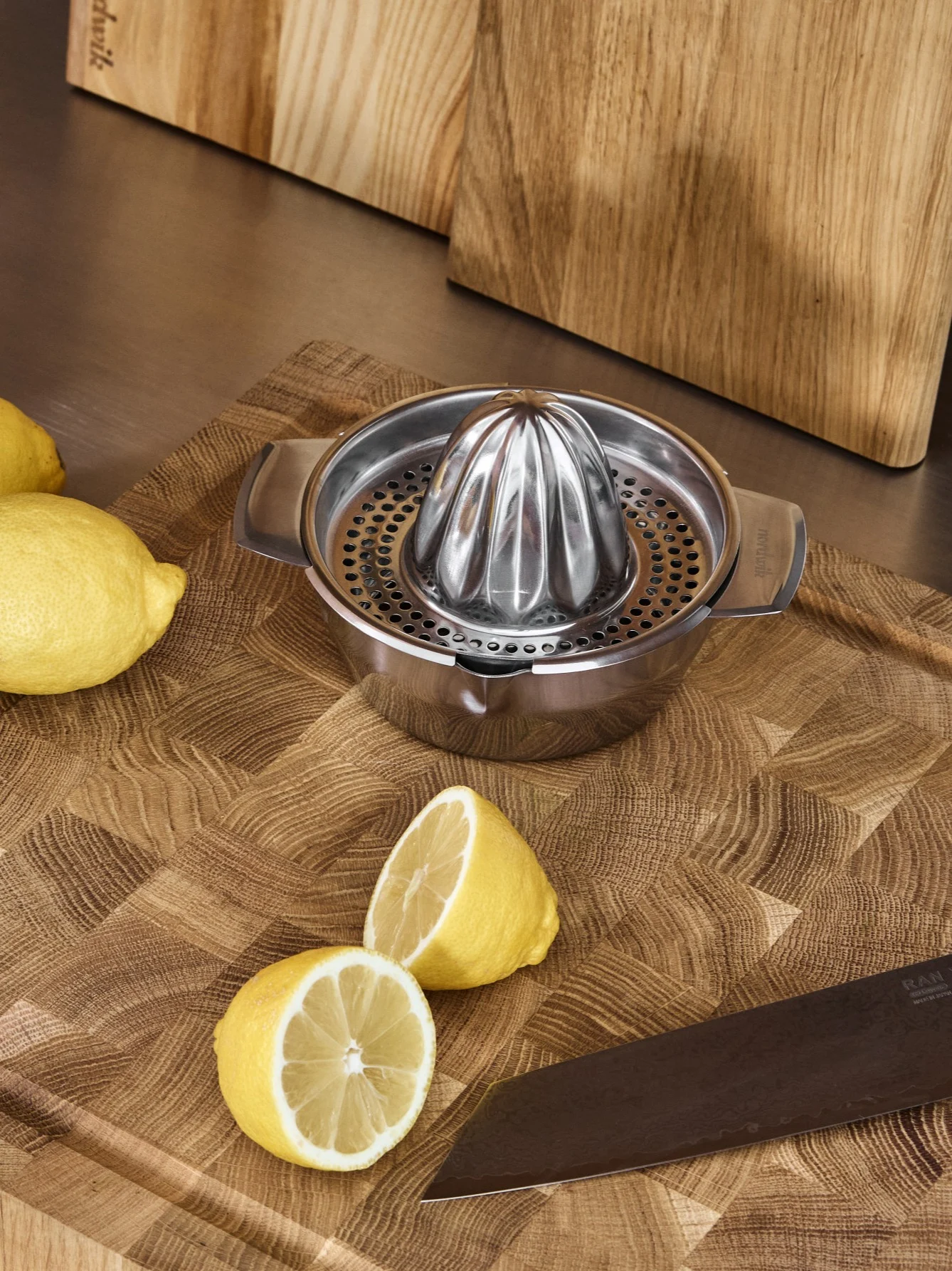 Spremiagrumi manuale in acciaio inossidabile, limoni interi e a metà, e un coltello su un tagliere di legno.