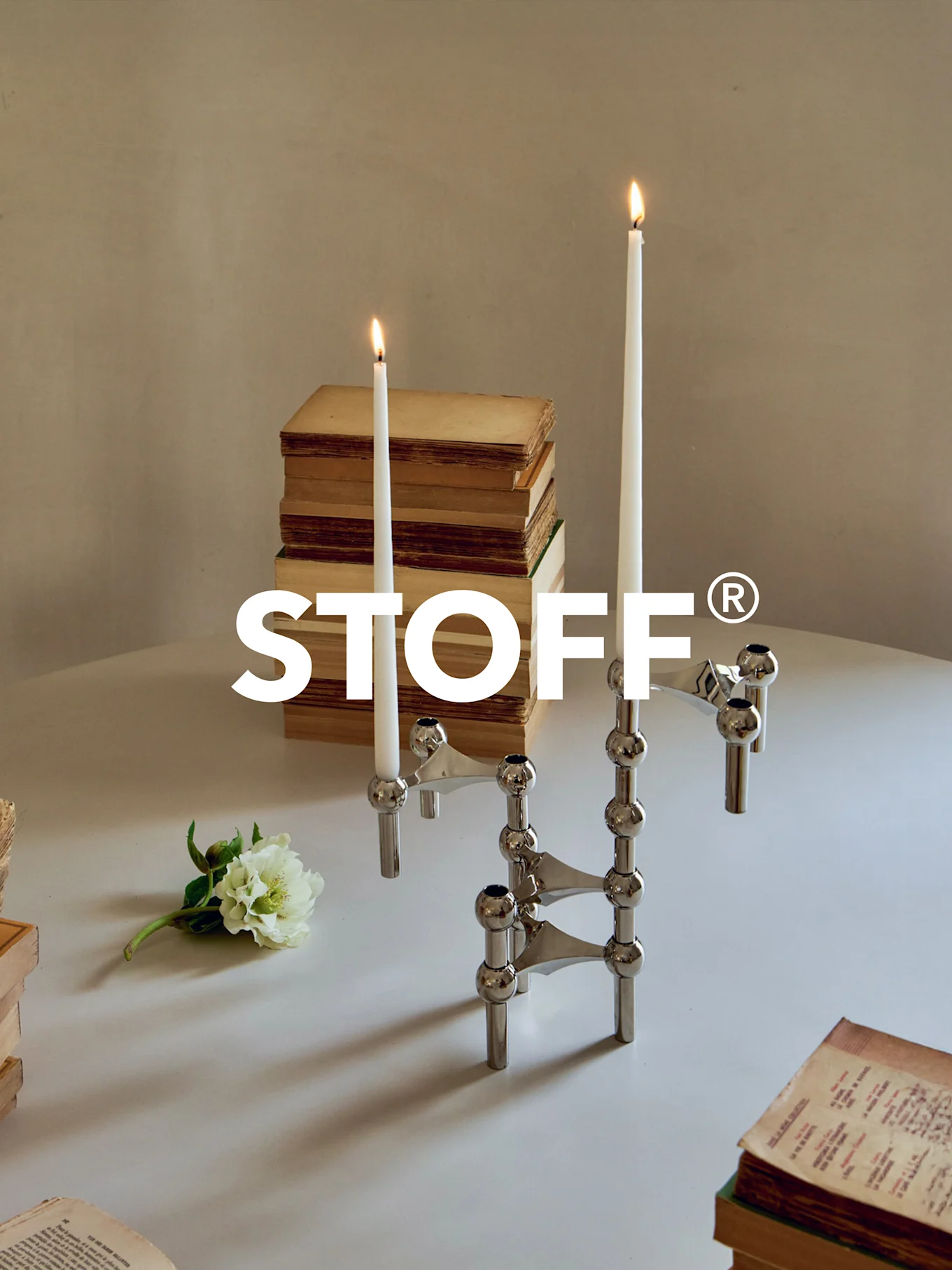 Candelabro modulare in cromo lucido con due candele accese, circondato da libri antichi e un fiore bianco su un tavolo. Logo STOFF®.