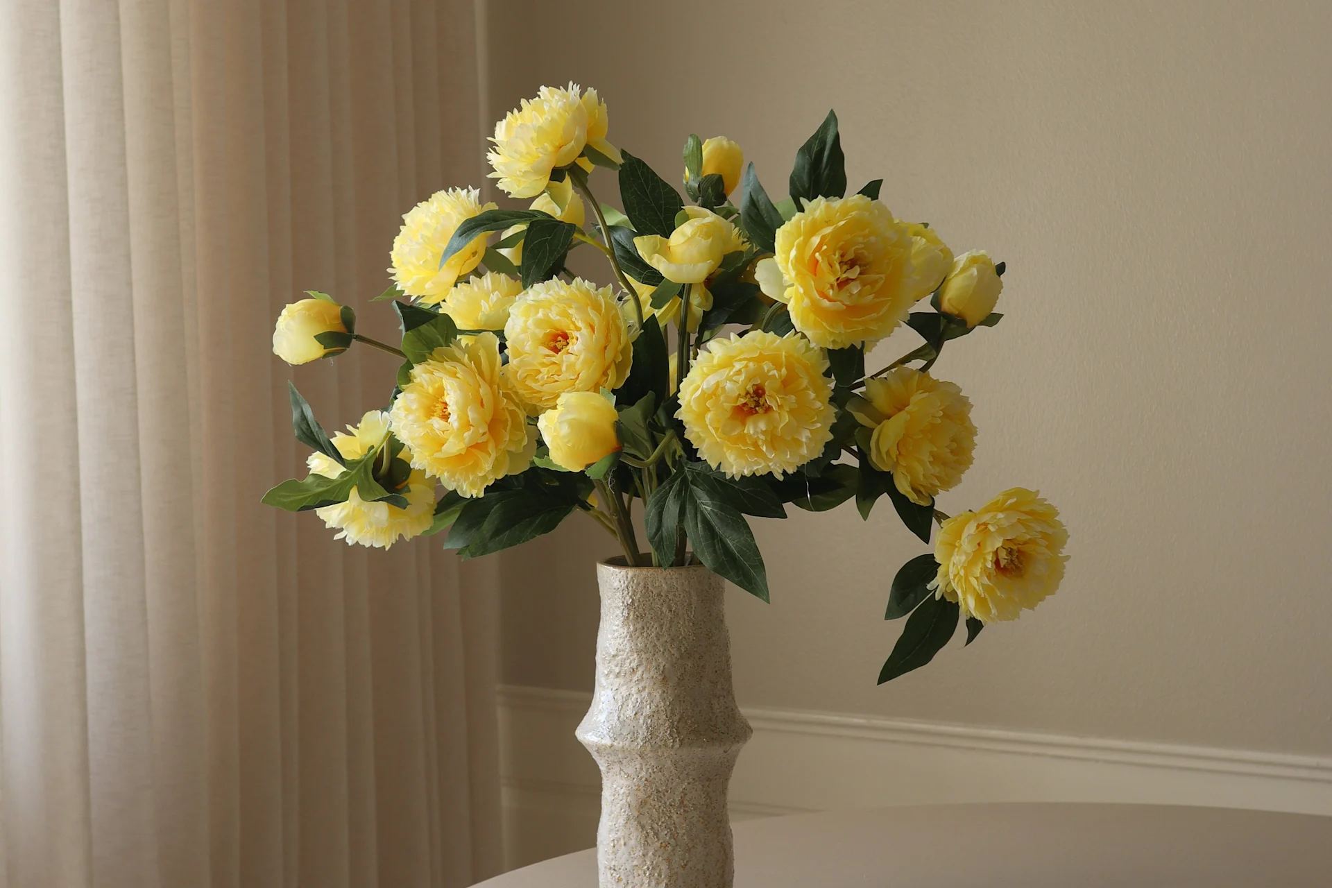 Un vaso chiaro e testurizzato contiene un vivace bouquet di fiori gialli simili a peonie e foglie verdi su uno sfondo chiaro.