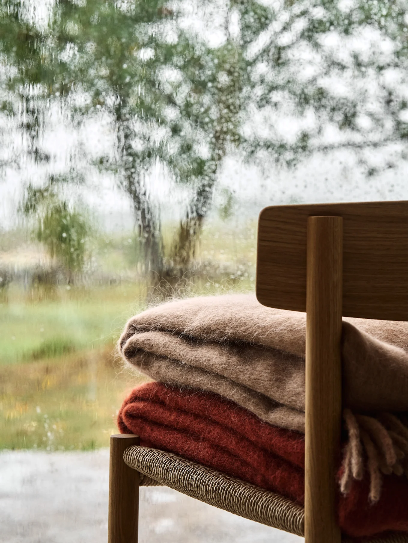 Scena accogliente con due soffici coperte su una sedia di legno, vista attraverso una finestra bagnata dalla pioggia con un paesaggio verde sfocato.