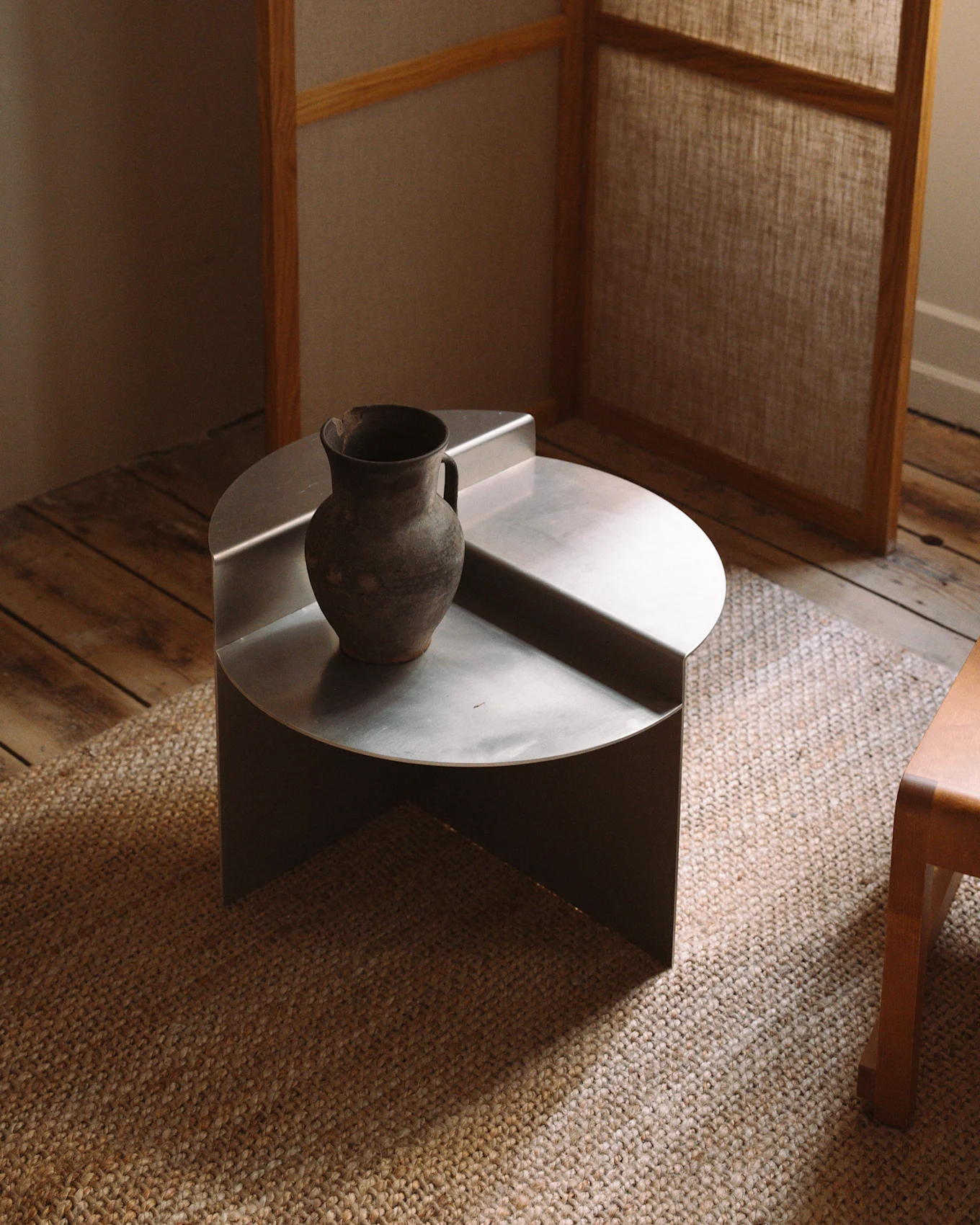 Un tavolino da caffè minimalista, basso e rotondo in metallo argentato con un vaso rustico in ceramica scura su un tappeto intrecciato.