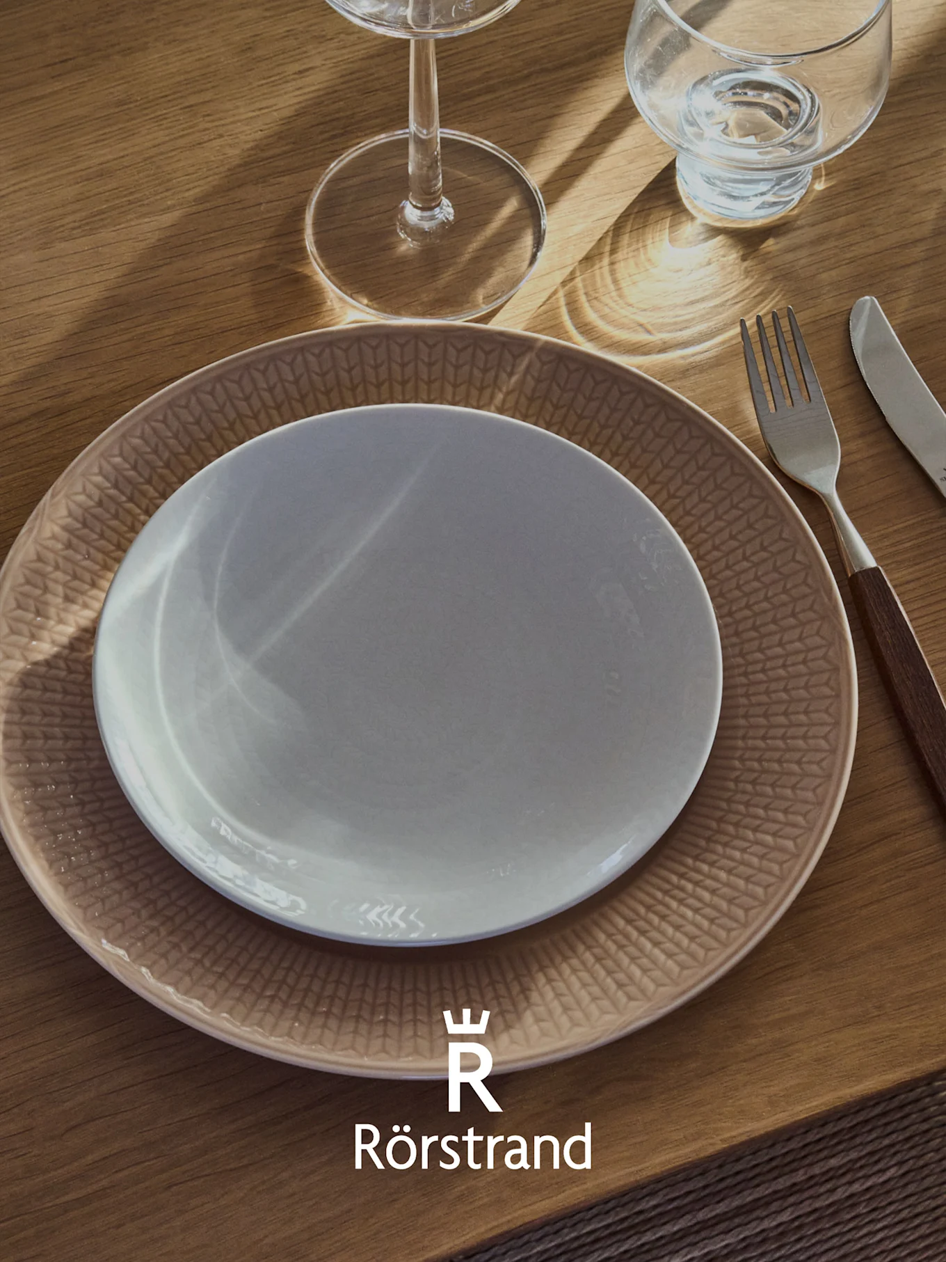 Un posto tavola con un piatto bianco su un piatto beige fantasia, una forchetta, un coltello e due bicchieri su un tavolo di legno. Logo Rörstrand.
