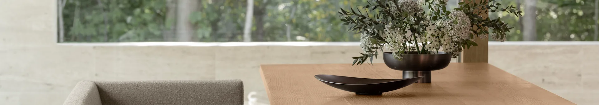 Tavolo da pranzo moderno in legno con una ciotola decorativa scura e un vaso di fiori bianchi e verdi, con una vista esterna verde sfocata attraverso una grande finestra.