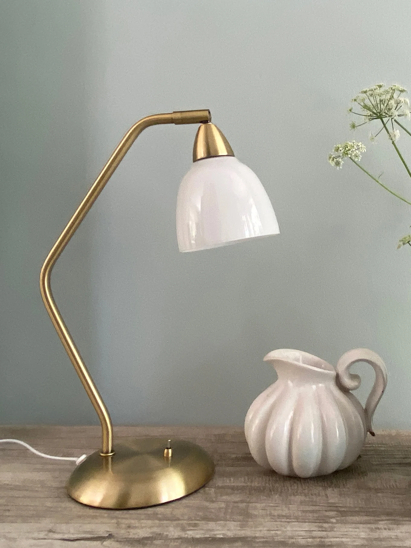 Lampada da tavolo in ottone con paralume in vetro bianco, accanto a brocca in ceramica bianca scanalata su legno.