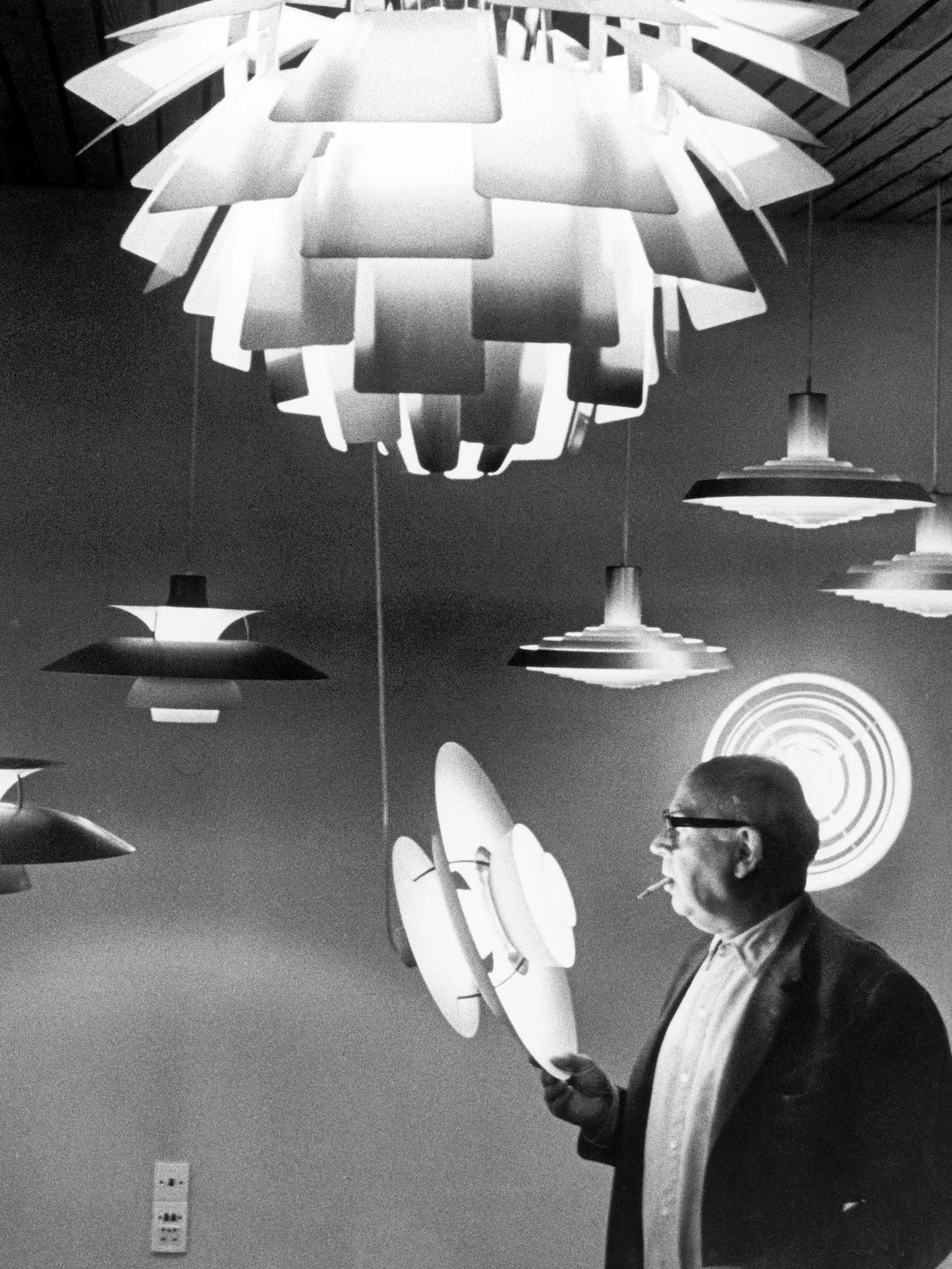 Uomo con occhiali e sigaretta ispeziona diverse lampade moderne a strati in bianco e nero.