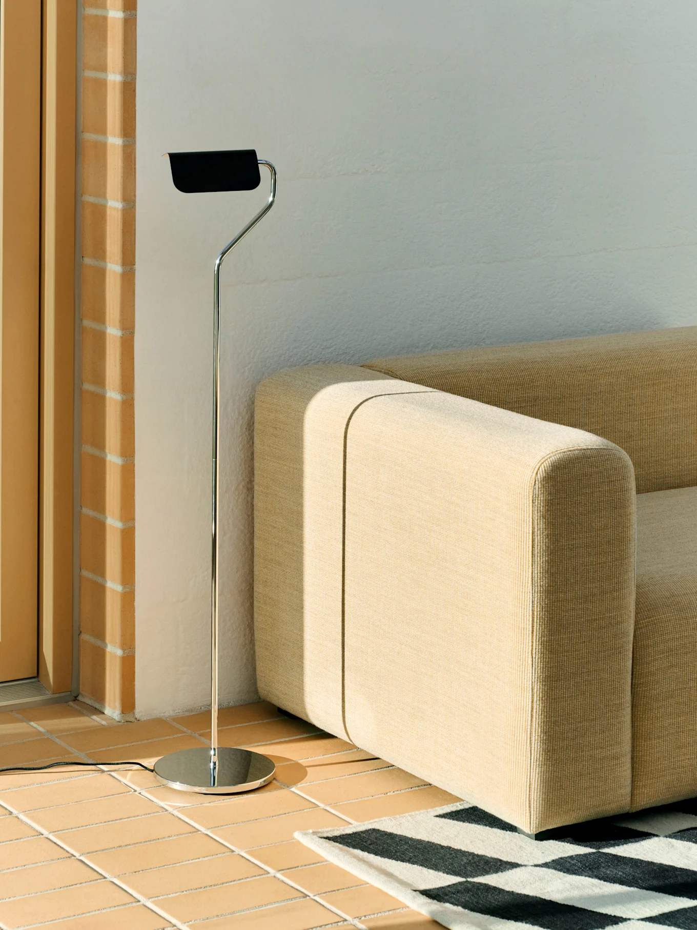 Una lampada da terra cromata con paralume nero accanto a un divano modulare beige e un tappeto geometrico bianco e nero su un pavimento piastrellato.