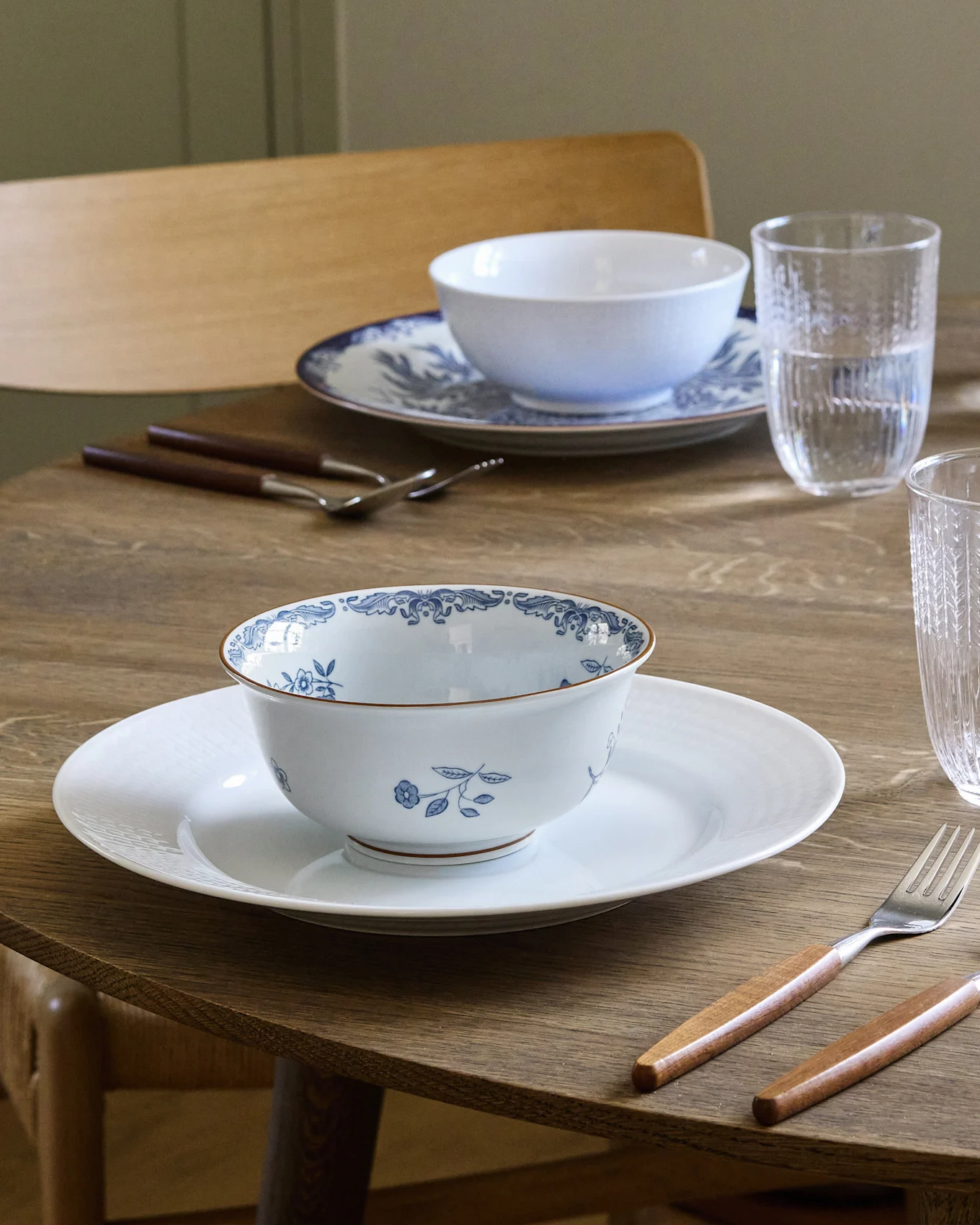 Tavolo da pranzo apparecchiato per due con ciotole a fantasia bianca e blu su piatti bianchi, posate con manico in legno e bicchieri d'acqua.