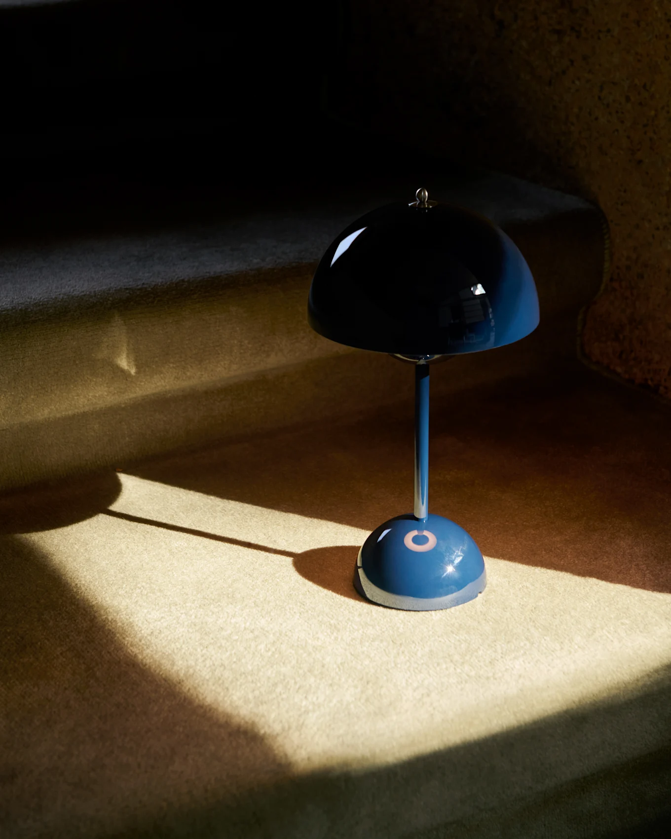 Una lampada da tavolo blu a forma di fungo con paralume riflettente su una superficie in moquette, parzialmente illuminata dal sole.