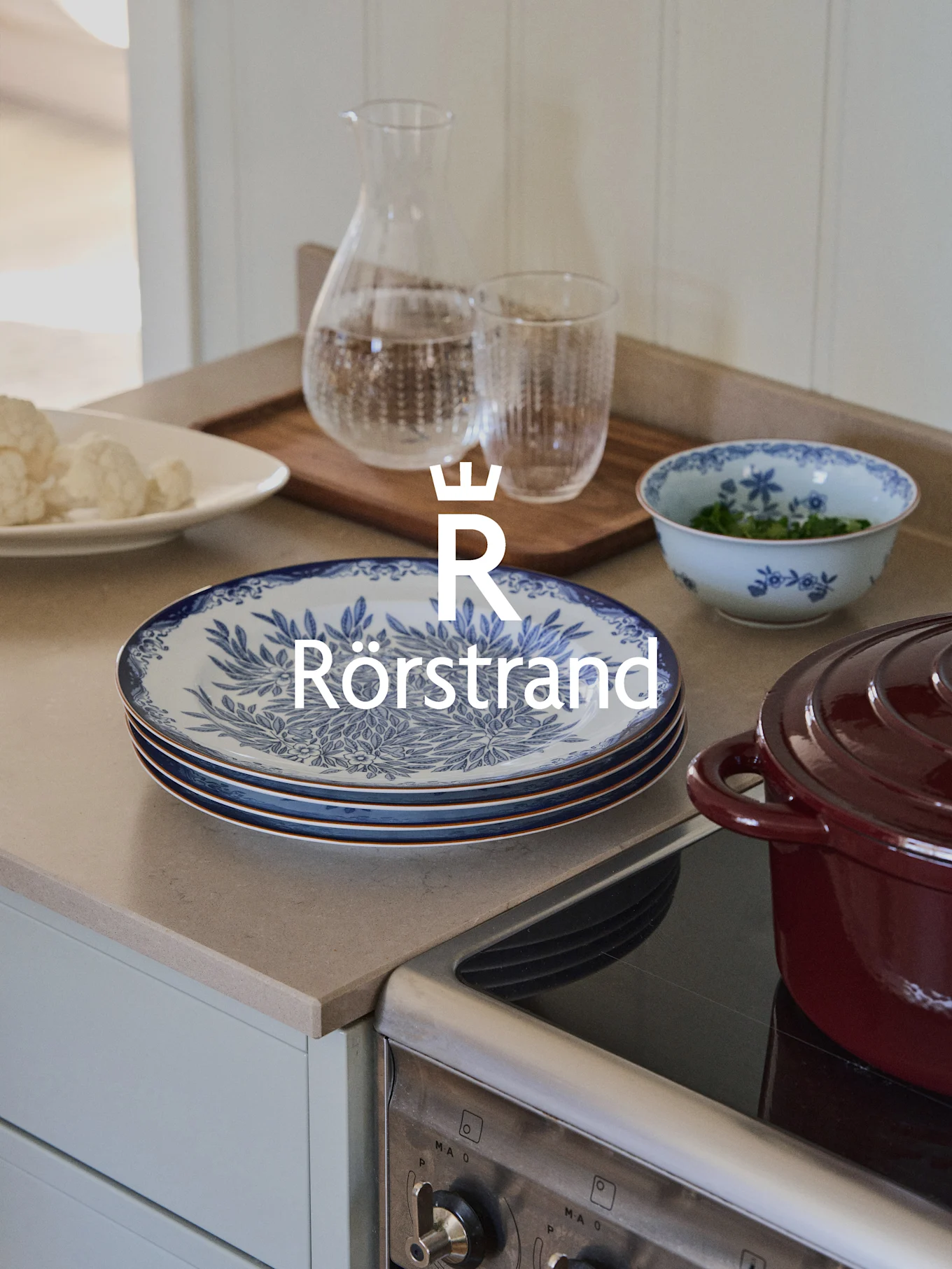 Un bancone da cucina con piatti Rörstrand, una caraffa e una ciotola con motivi floreali blu, accanto a una pentola rossa su un fornello.