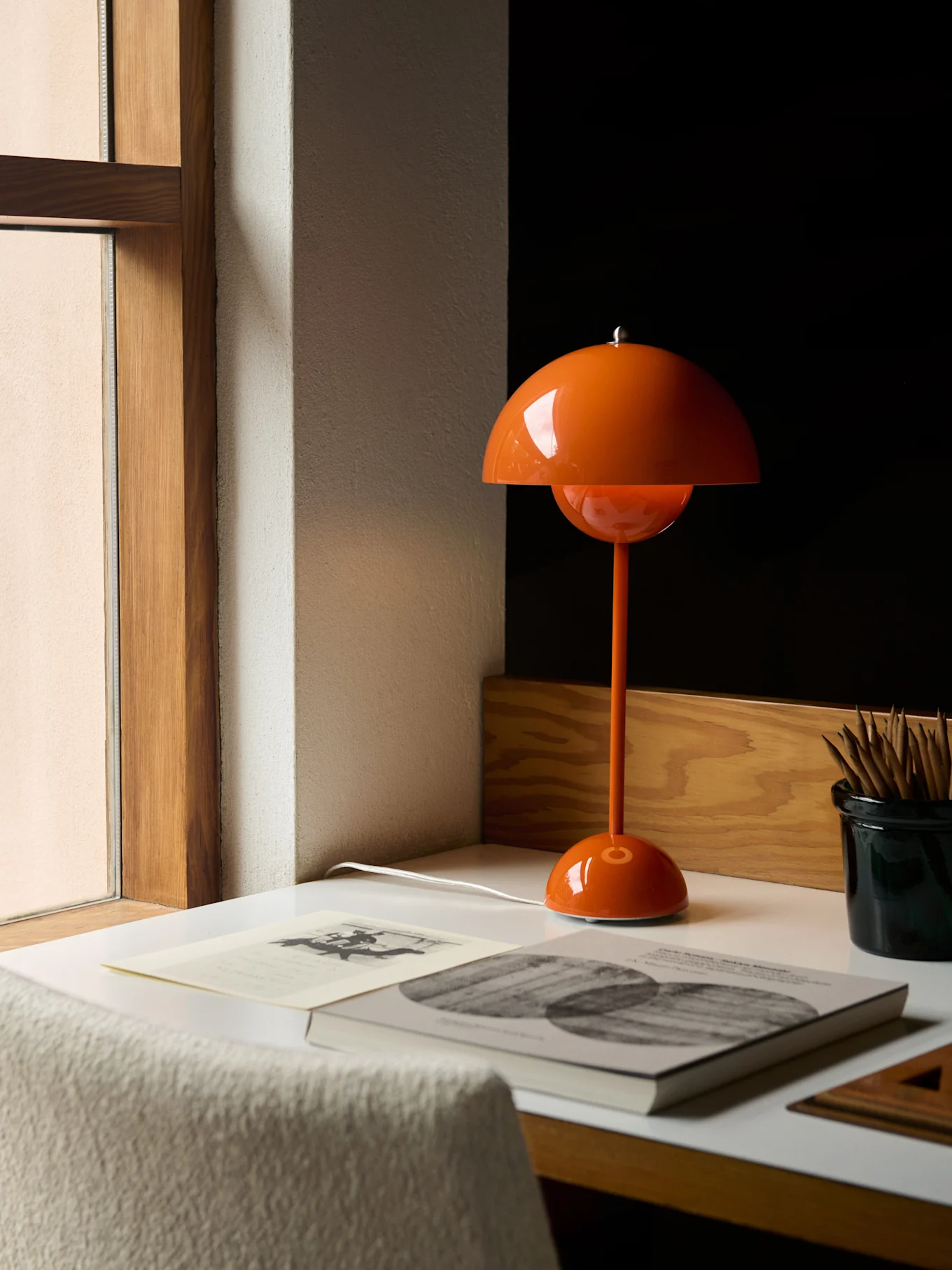 Una lampada da tavolo arancione a forma di fungo su una scrivania bianca accanto a una finestra, con libri e matite.