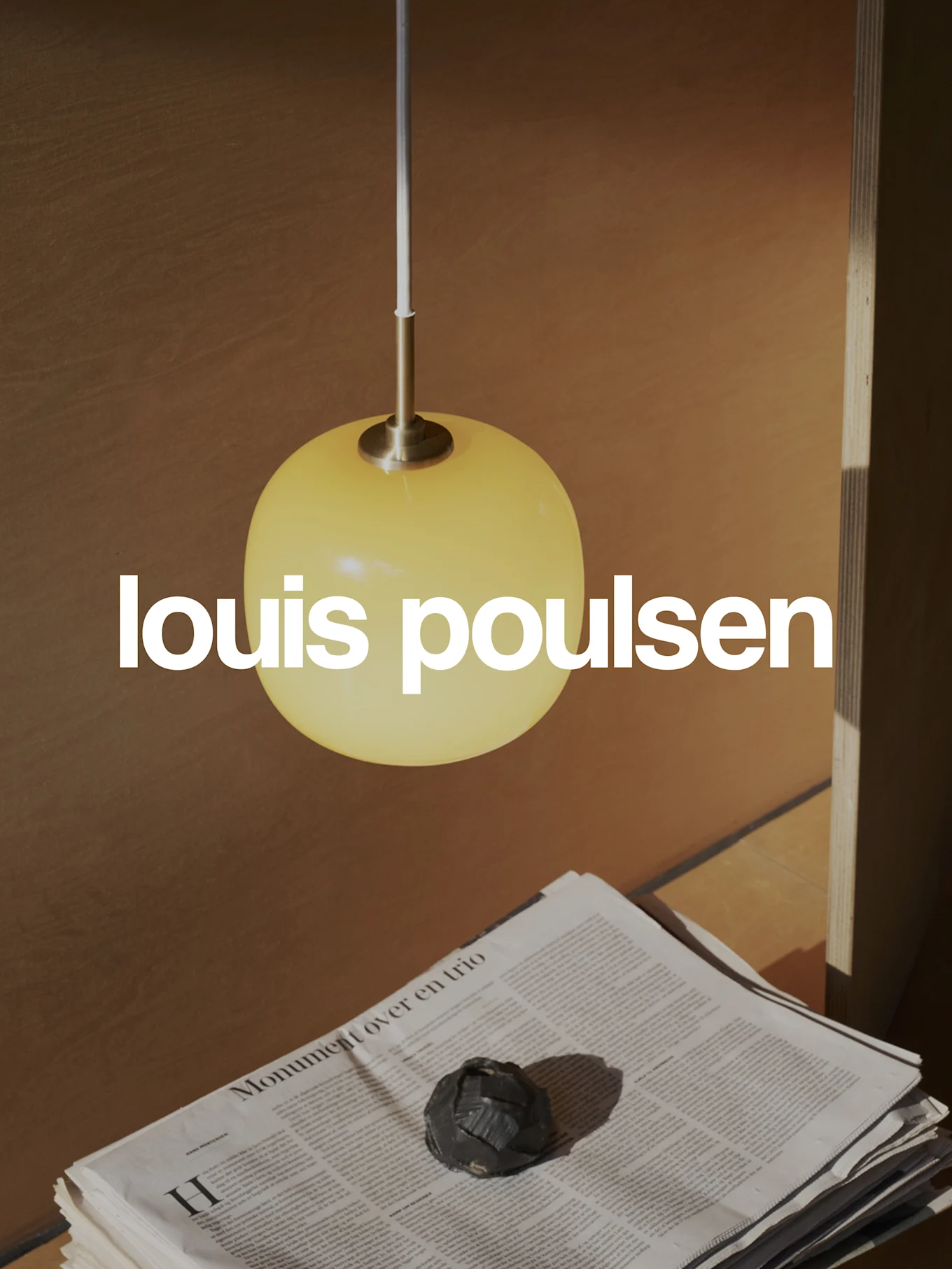 Logo Louis Poulsen sovrapposto a una scena calda con una lampada a sospensione sferica gialla, giornali impilati e un oggetto decorativo scuro.