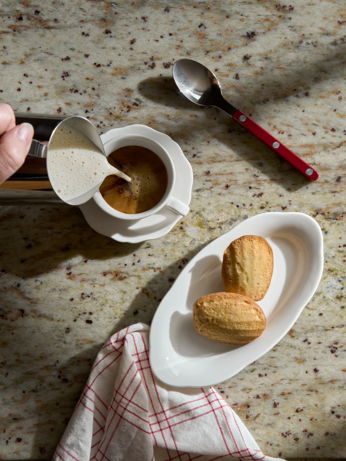 Mano che versa latte schiumato in una tazza di caffè accanto a madeleine su un ripiano in granito.