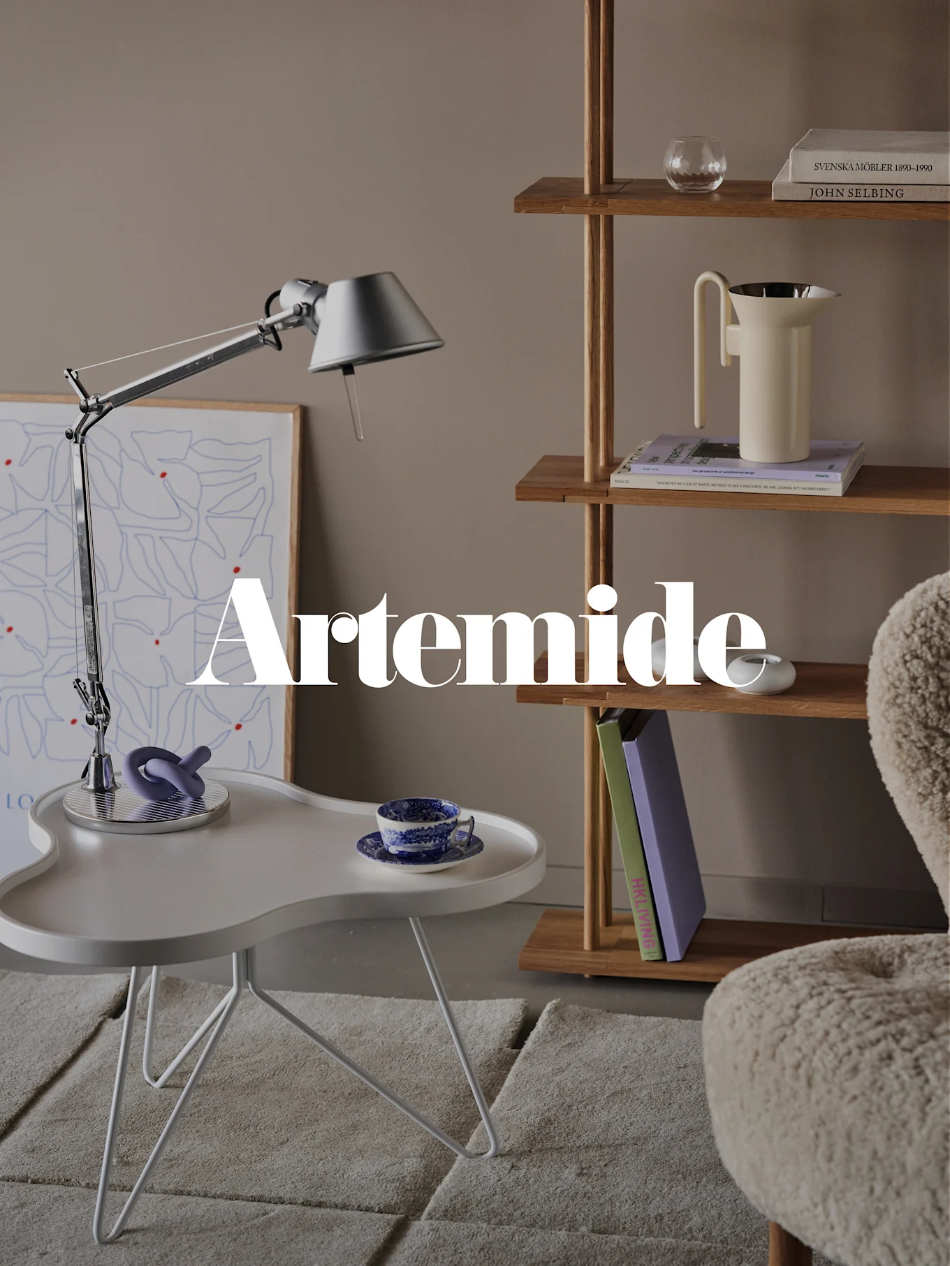 Un soggiorno moderno con lampada da scrivania argentata, tavolino bianco, libreria in legno e poltrona color crema. Sovrapposizione testo: 'Artemide'.