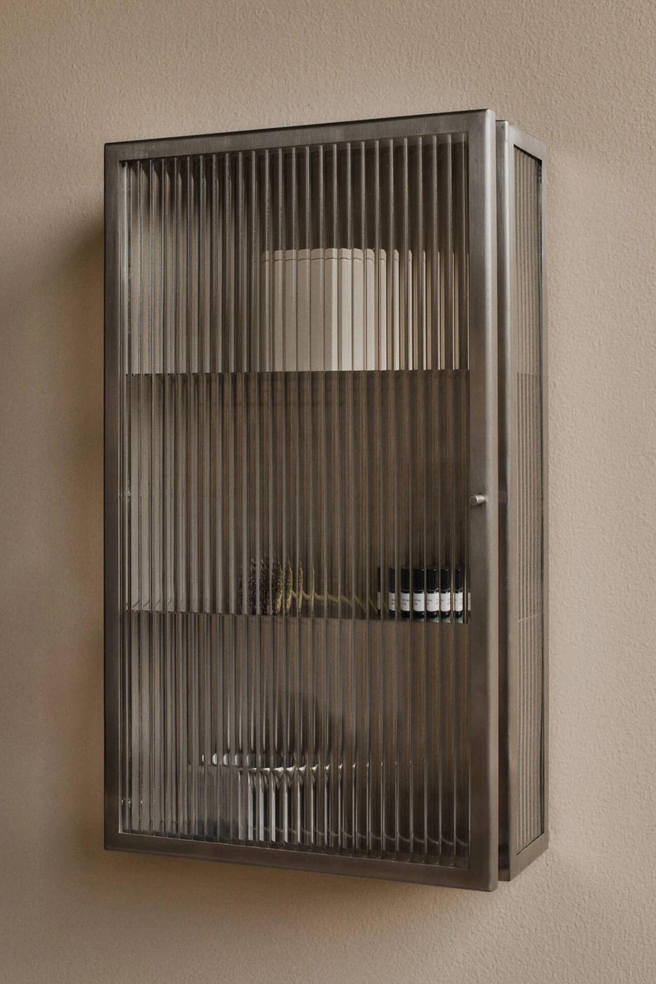 Mobiletto in metallo scuro a parete con ante in vetro scanalato su una parete beige. Contiene mensole con un cilindro bianco, piante essiccate e piccole bottiglie scure.