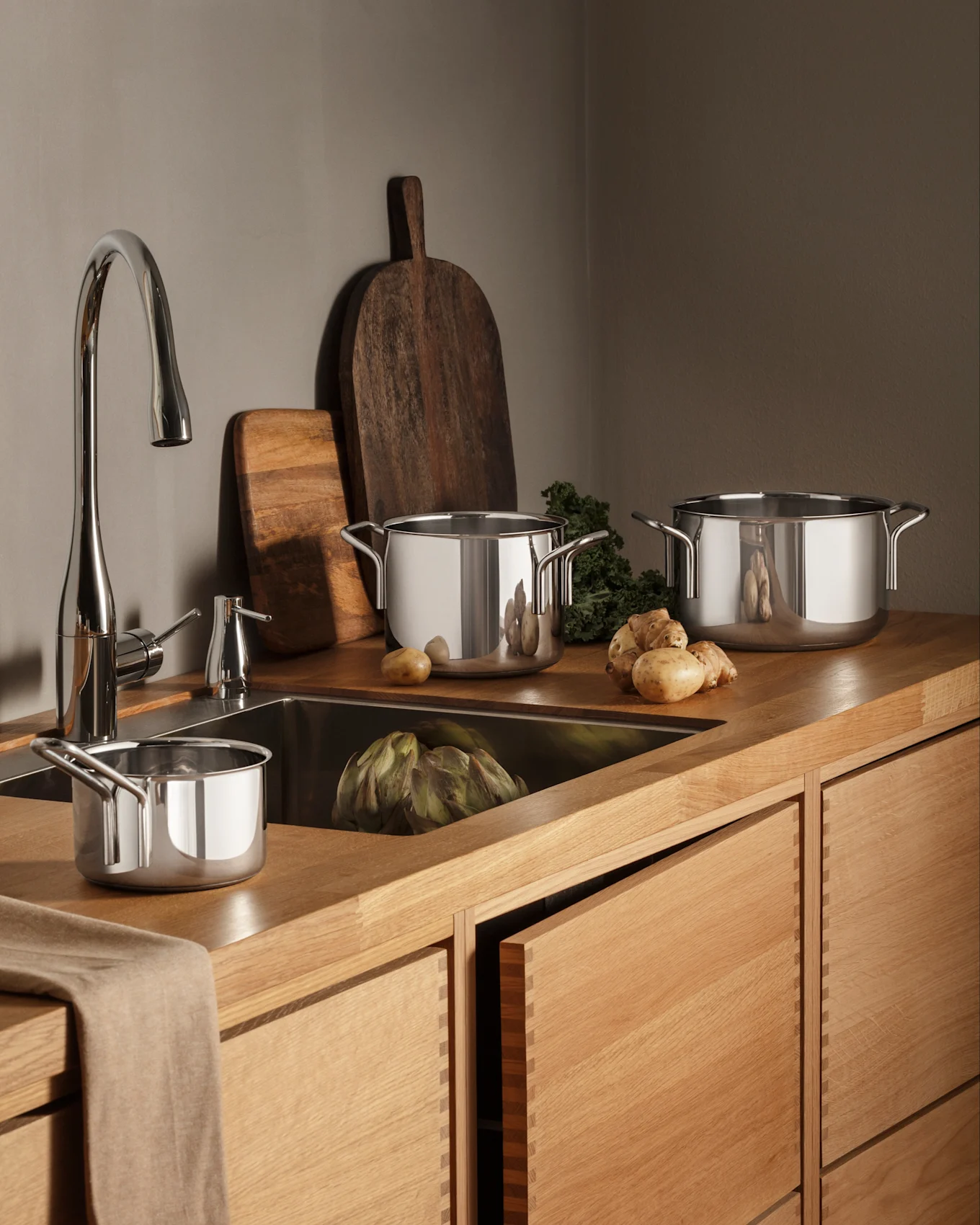 Una cucina moderna con banconi e mobili in legno, un rubinetto cromato e pentole in acciaio inossidabile, prodotti freschi tra cui carciofi e patate.