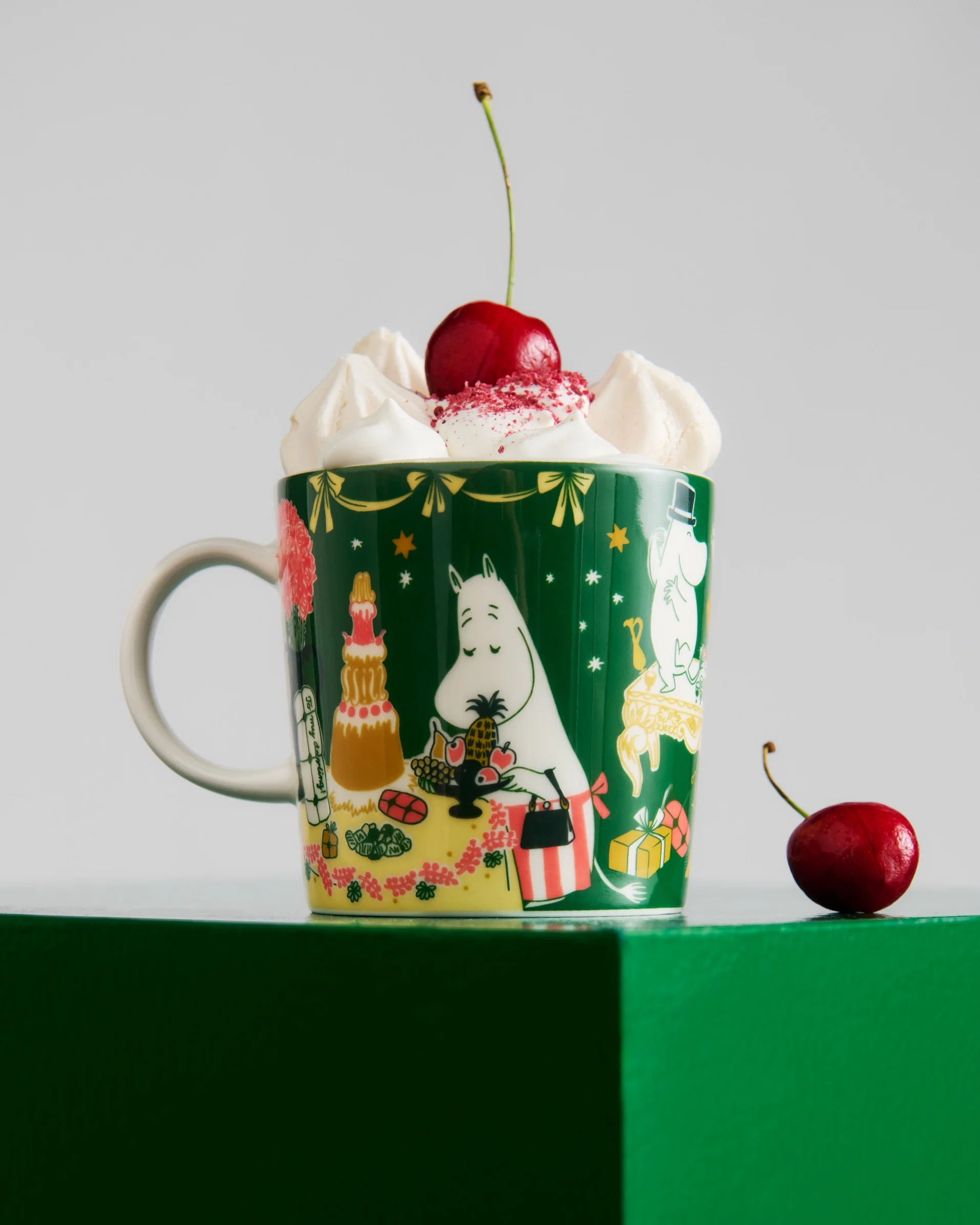 Tazza verde scuro con personaggi Moomin, guarnita con panna montata, granella rossa e una ciliegia. Un'altra ciliegia è sulla superficie verde.