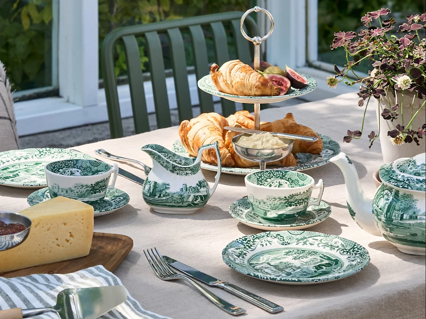 Un tavolo all'aperto apparecchiato con stoviglie a fantasia verde e bianca, croissant, formaggio e una teiera, in un giardino.
