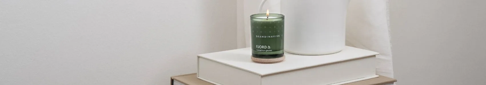 Un libro intitolato 'NOTES FROM THE FELLS' su una coperta grigia accanto a due candele in stile scandinavo, una nera e accesa, una marrone con coperchio in legno.