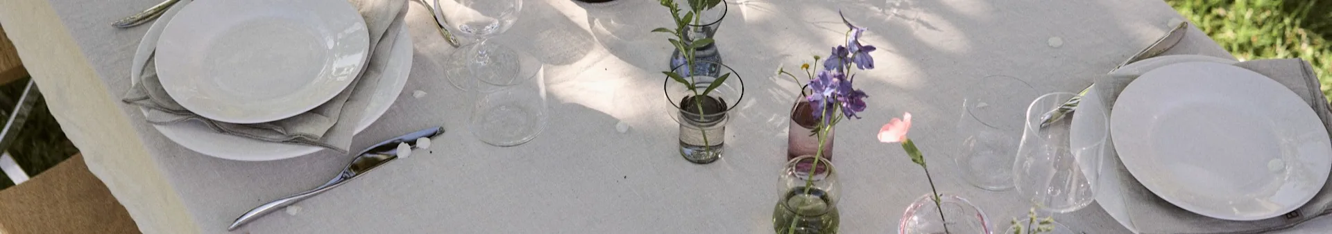 Tavolo all'aperto apparecchiato con piatti bianchi, posate e piccoli vasi con fiori viola, rosa e bianchi su una tovaglia di lino chiaro.