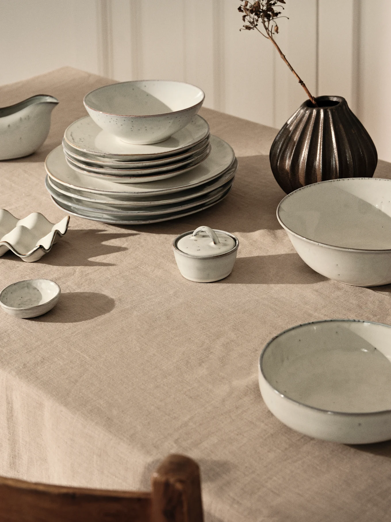 Servizio ceramica grigio chiaro, piatti e ciotole impilati su tovaglia lino beige, vaso scuro e ramo secco.