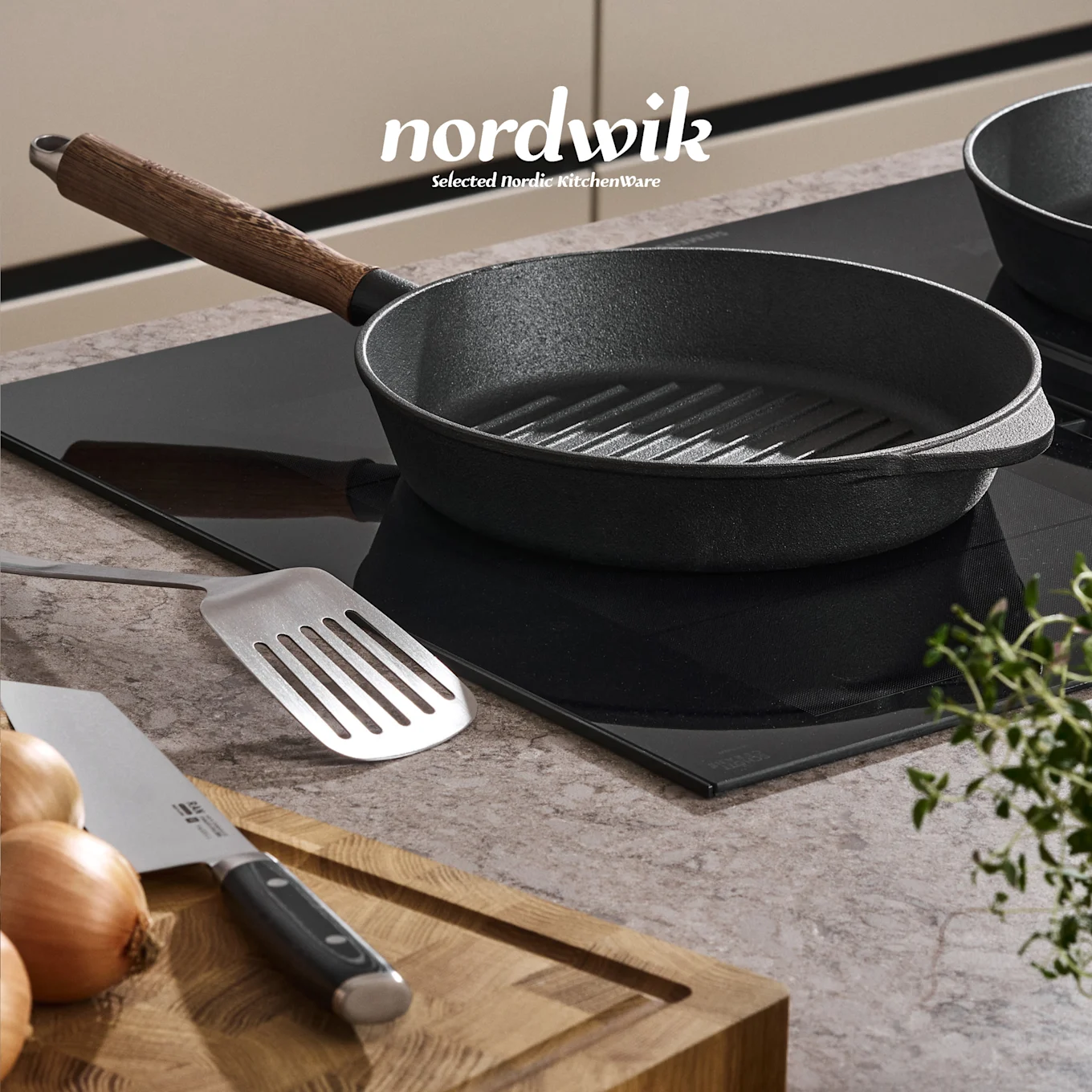 Una griglia in ghisa nera su un piano cottura a induzione, con una spatola, cipolle, coltello e timo su un tagliere in legno. Logo Nordwik visibile.