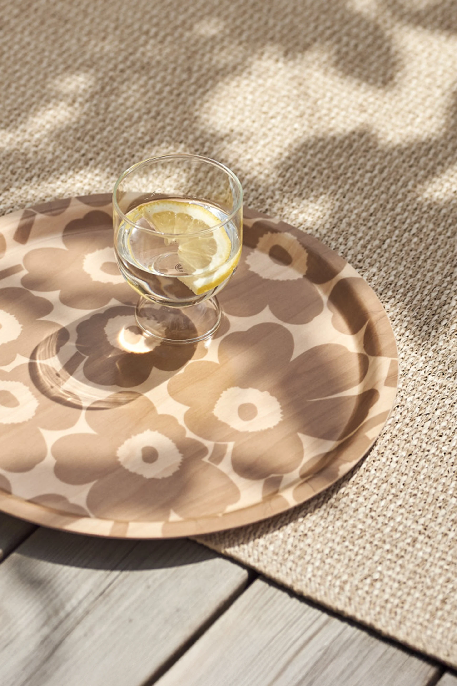 Bicchiere d'acqua con fetta di limone su un vassoio rotondo decorato, posato su un tappeto intrecciato beige e un ponte di legno con luce solare a chiazze.