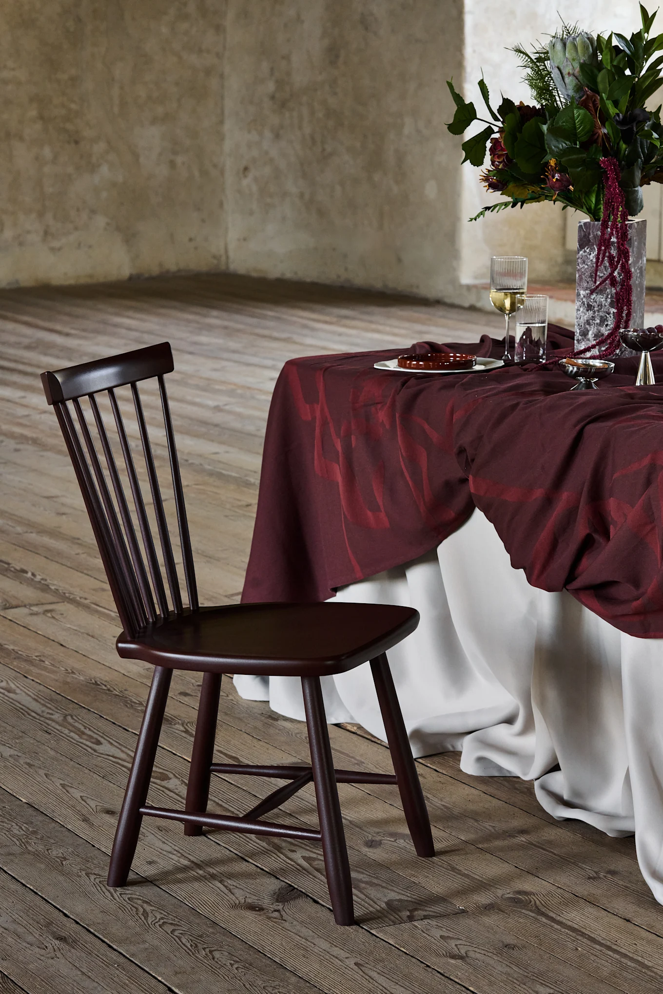 Sedia in legno marrone scuro con schienale a doghe, accanto a un tavolo drappeggiato in bordeaux con composizione floreale, su pavimento in legno.