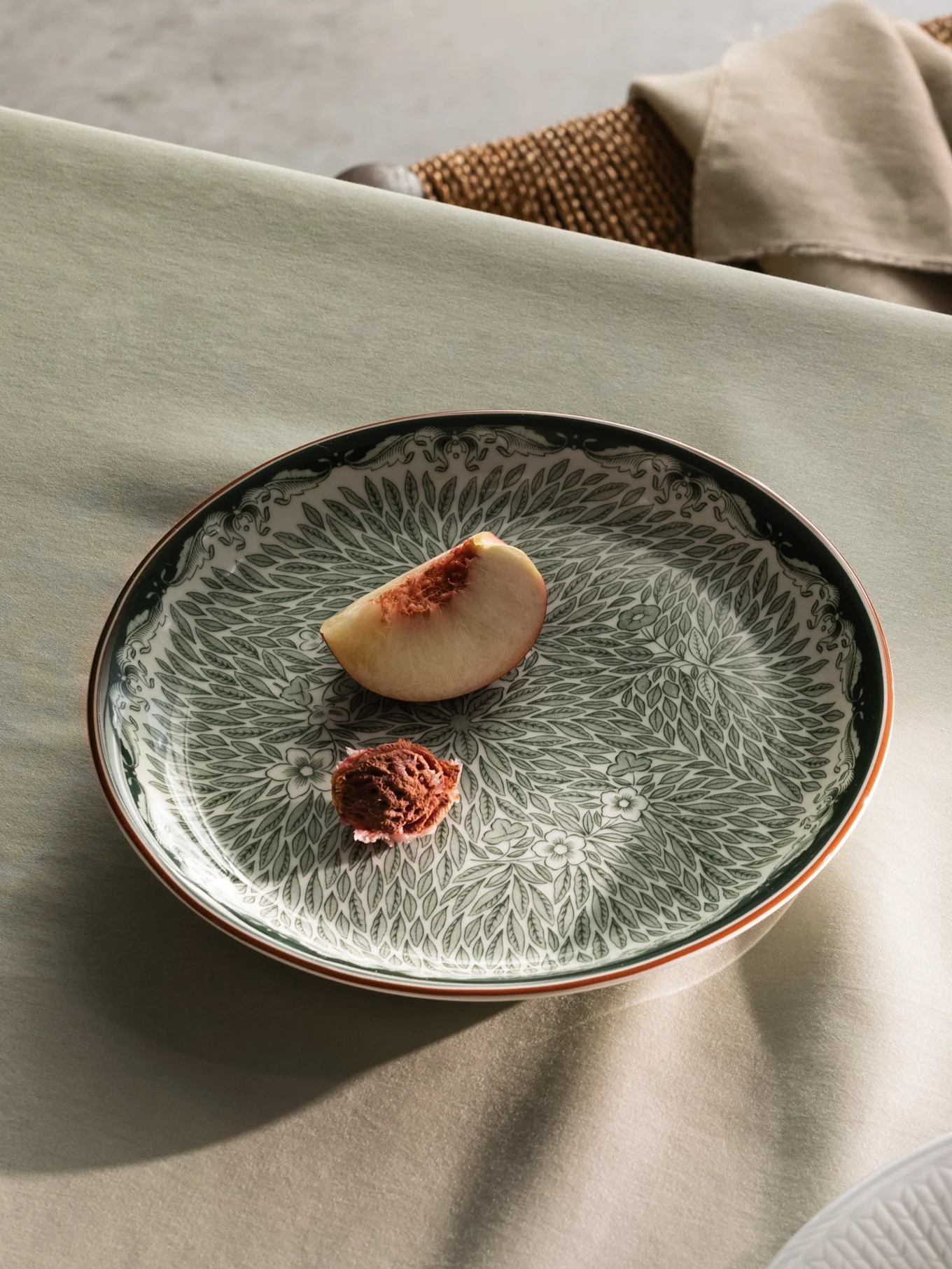 Un tavolo apparecchiato con un piatto verde decorato che contiene una fetta di pesca, e vista parziale di un piatto bianco con crema e fette di pesca.