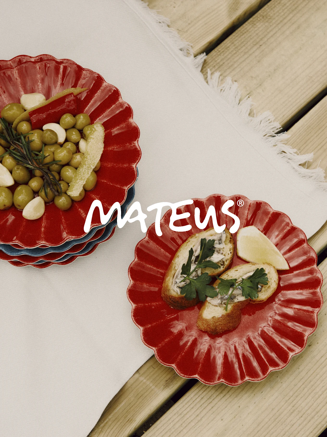 Piatti rossi smerlati con olive, aglio, rosmarino e pane con acciughe su tavolo in legno. Marchio MATEUS.