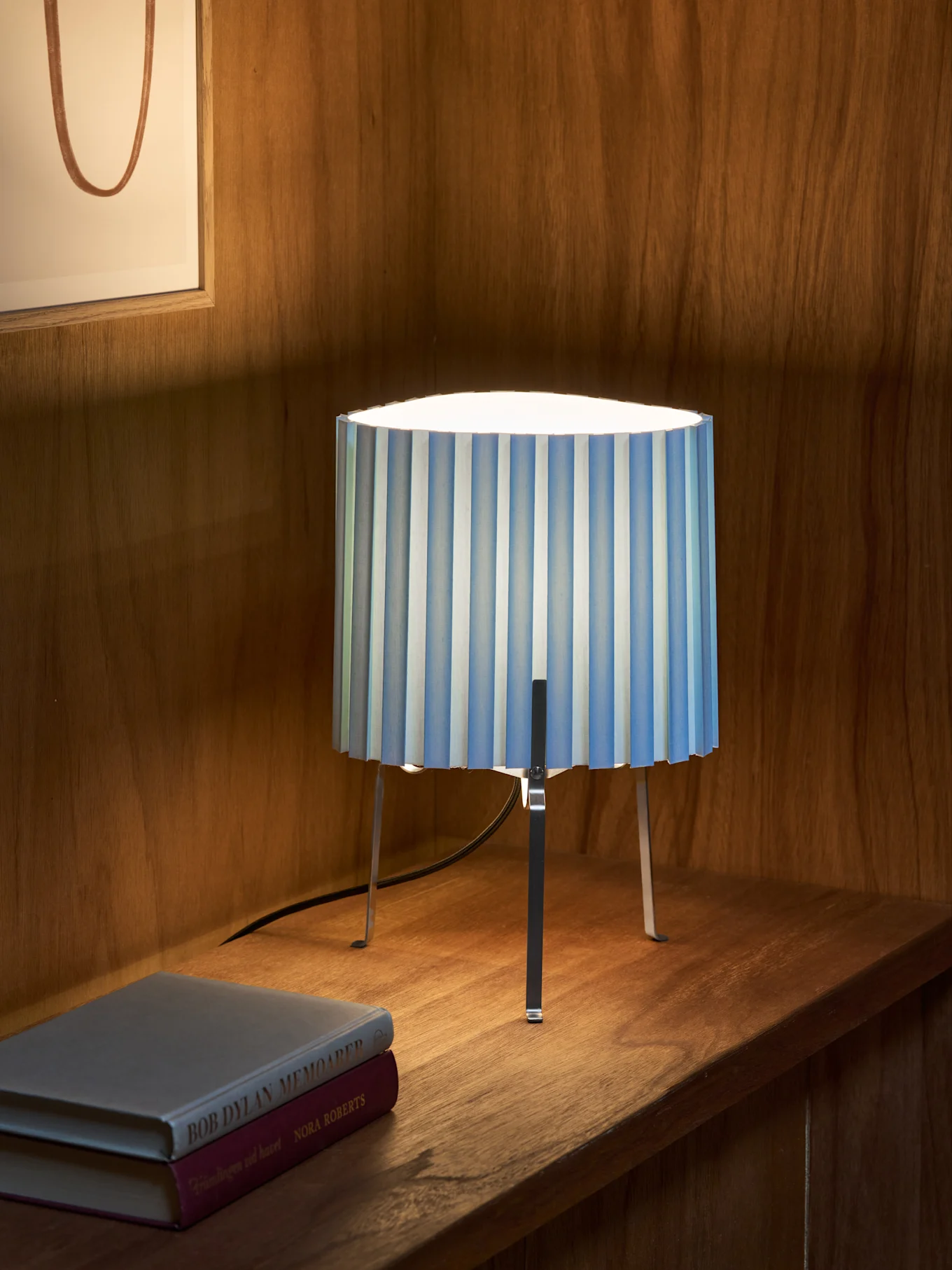 Una lampada da tavolo plissettata a righe blu e bianche con tre gambe metalliche su una mensola di legno accanto a libri. Opera d'arte incorniciata.