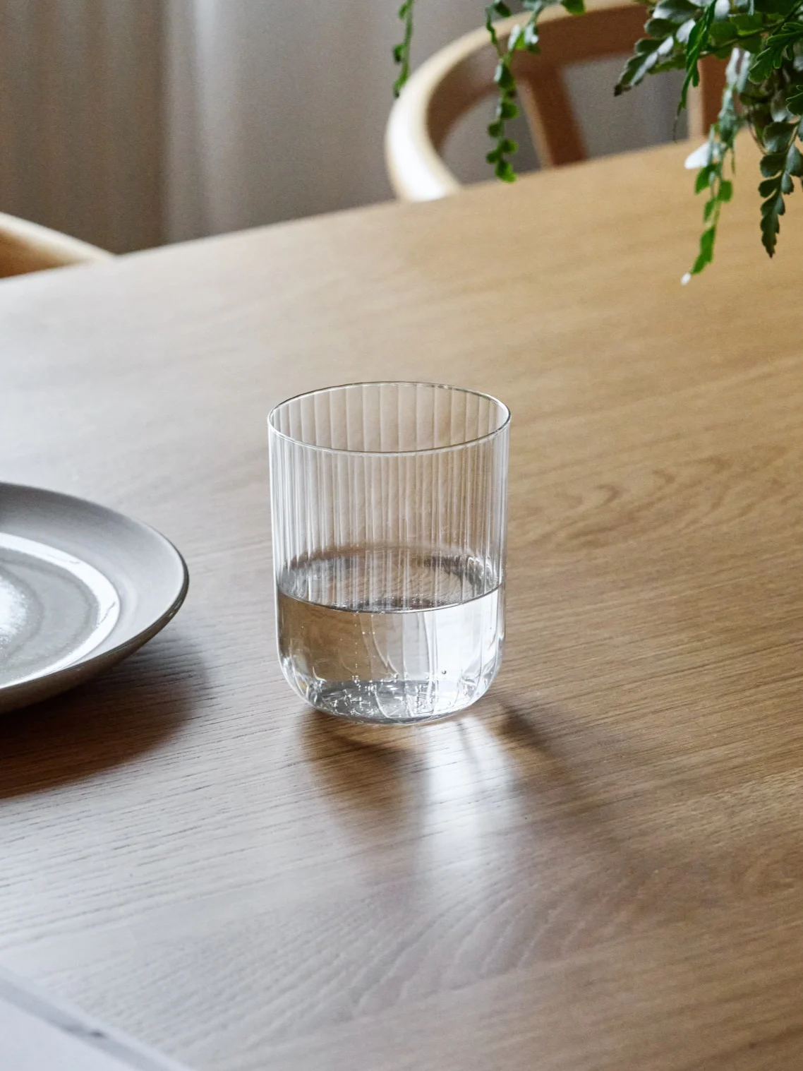 Un bicchiere di vetro a coste, mezzo pieno d'acqua, appoggiato su un tavolo di legno accanto a un piatto di ceramica.