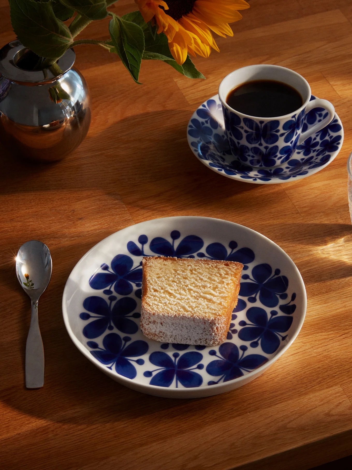 Una fetta di torta su un piatto floreale blu e bianco, con una tazza di caffè, un bicchiere d'acqua, un cucchiaio e un girasole in un vaso d'argento su un tavolo di legno soleggiato.