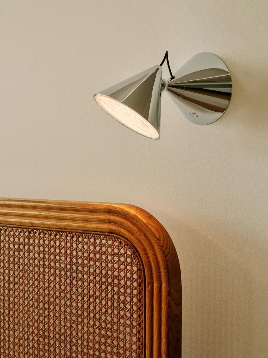 Lampada da parete cromata moderna con paralume conico che illumina una parete chiara sopra una testiera in canna intrecciata e legno.