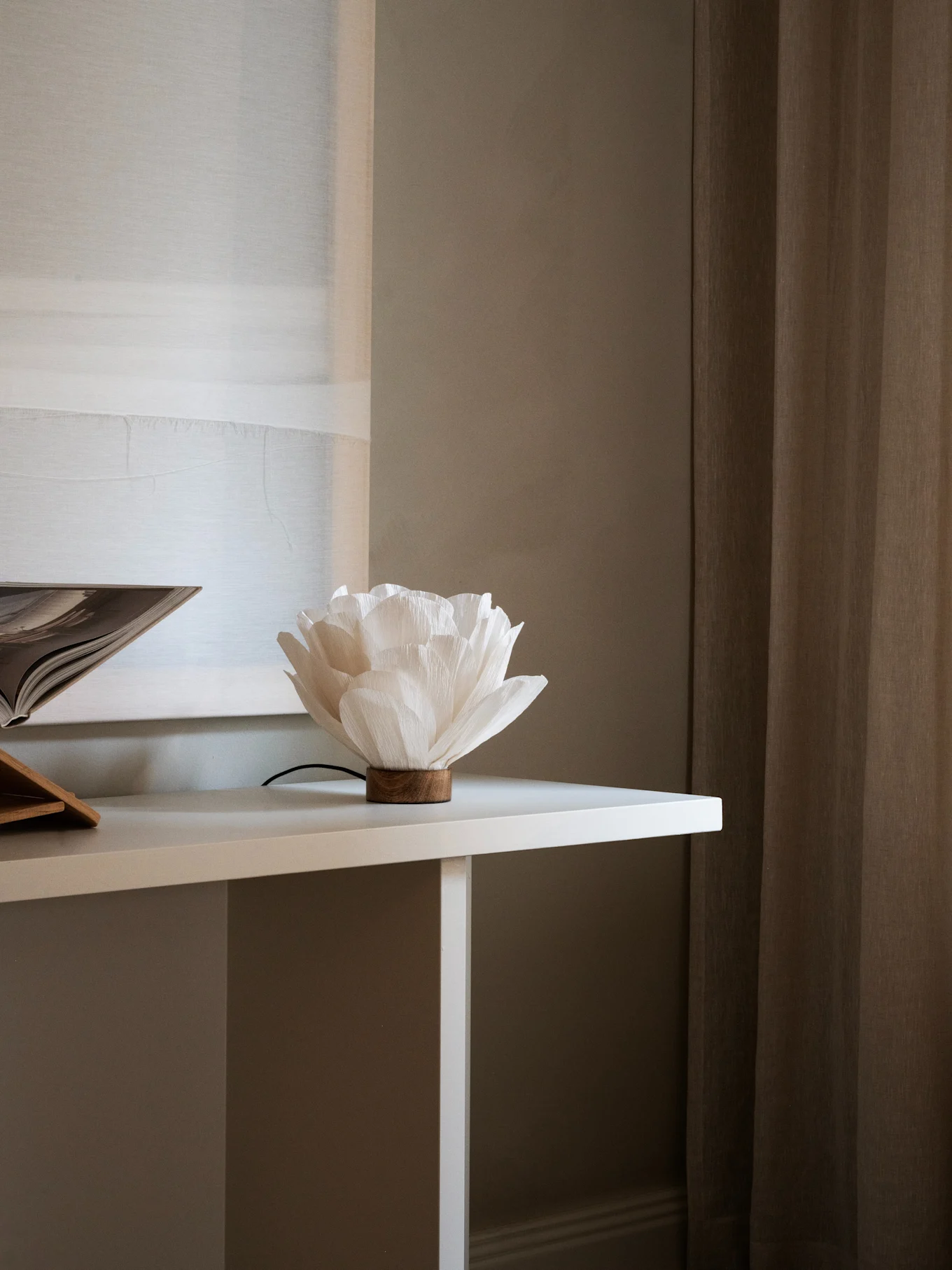 Lampada in carta bianca a forma di fiore con base in legno su un tavolo bianco, accanto a un libro aperto. Tende marroni sullo sfondo.