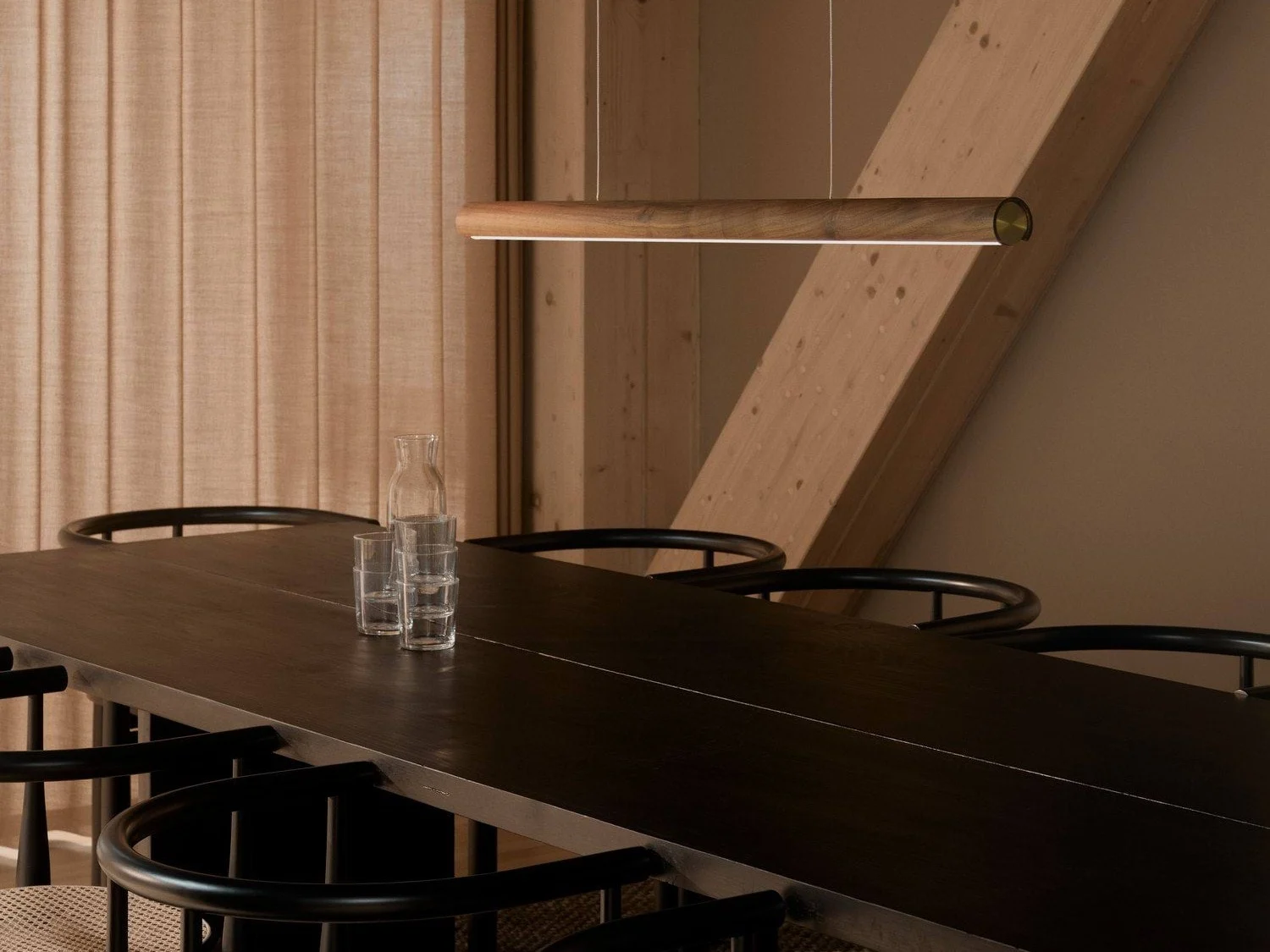 Una sala da pranzo moderna con un lungo tavolo in legno scuro, sedie nere e un'elegante lampada a sospensione in legno. Una caraffa di vetro e tre bicchieri sono sul tavolo.