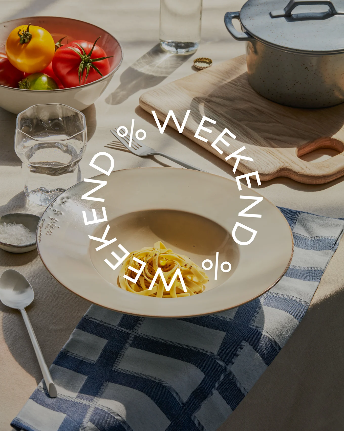 Tavolo con pasta, una ciotola di pomodori freschi, bicchiere d'acqua e pentola. Il testo 'WEEKEND % WEEKEND %' è sovrapposto.