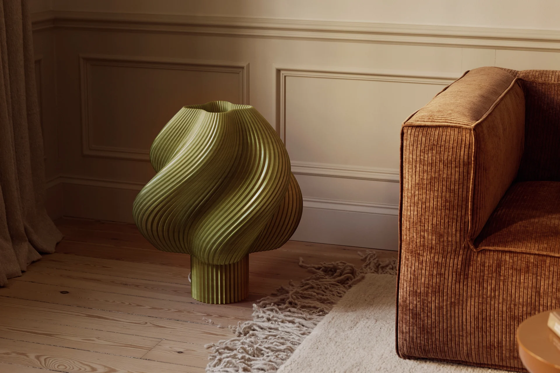 Un oggetto scultoreo verde oliva scanalato su un pavimento in legno accanto a un divano in velluto a coste marrone e un tappeto testurizzato in una stanza con pareti modanate.