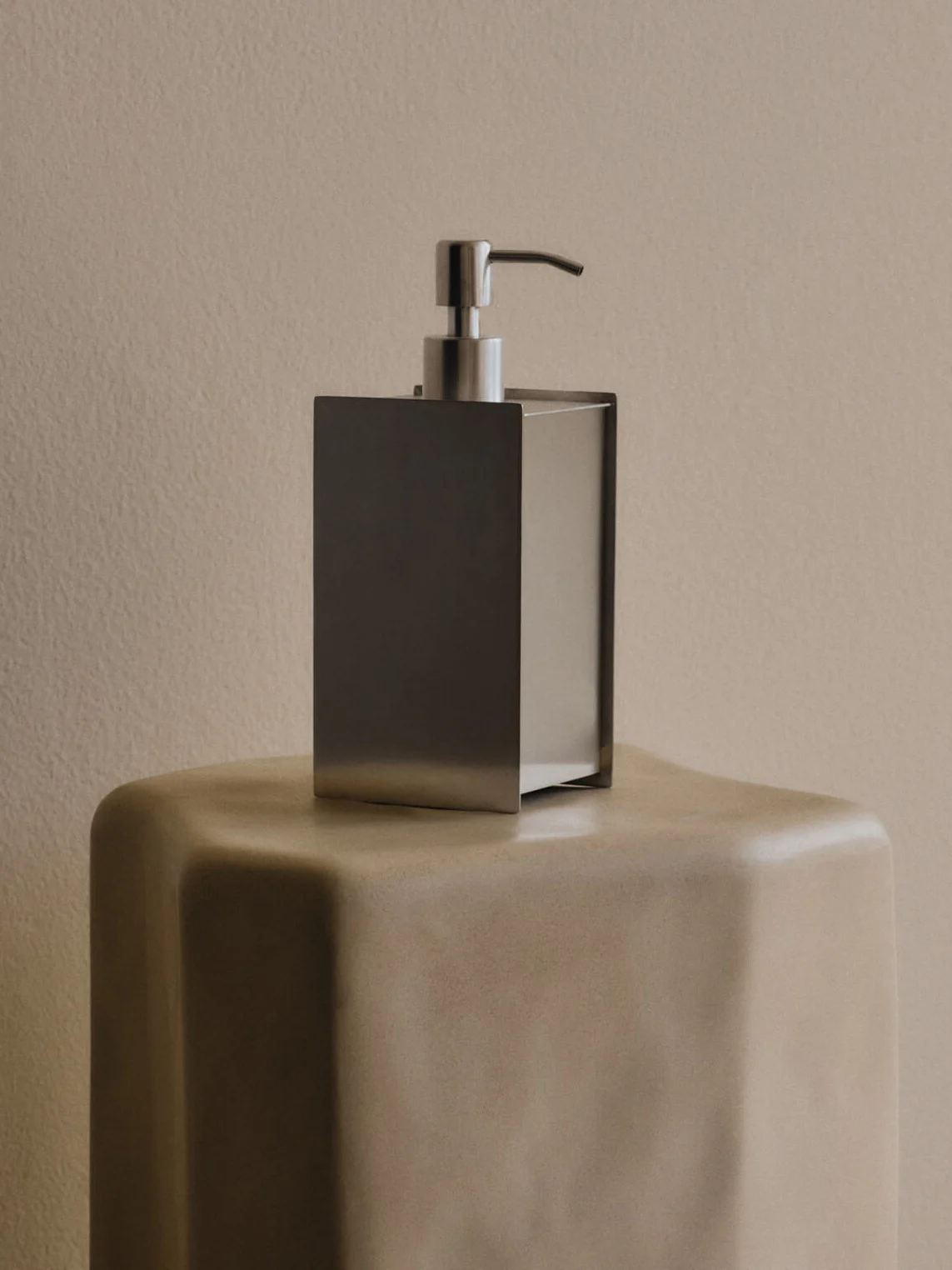 Elegante dispenser di sapone rettangolare in acciaio inossidabile su un piedistallo beige.