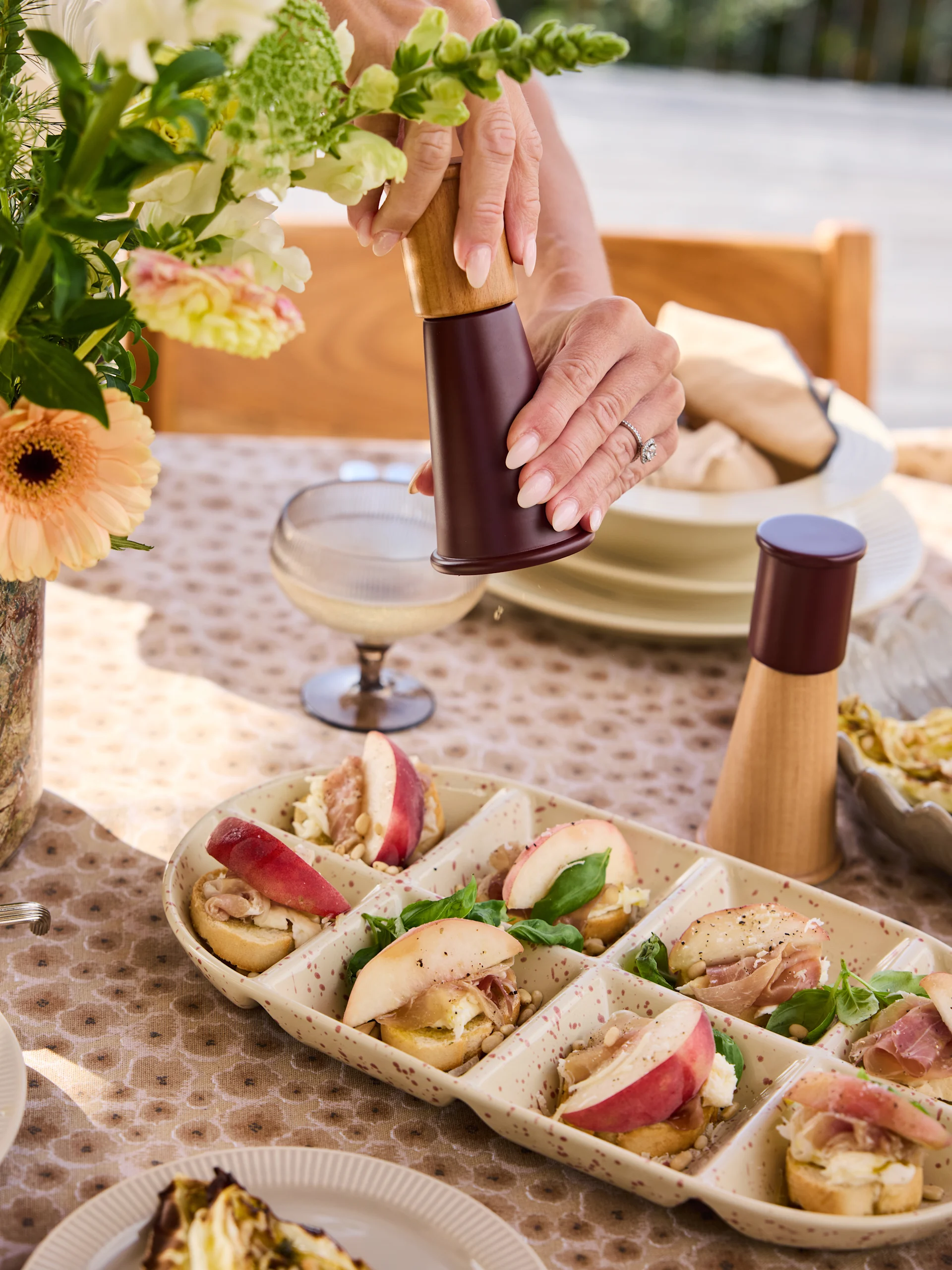 Mani che macinano pepe su un piatto di crostini con pesche, prosciutto, formaggio, pinoli, basilico.