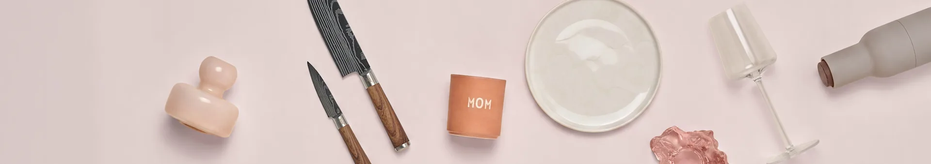 Ceramica rosa, due coltelli, tazza 'MOM', piatto bianco, vetro rosa, calice e bottiglia grigia su superficie rosa.