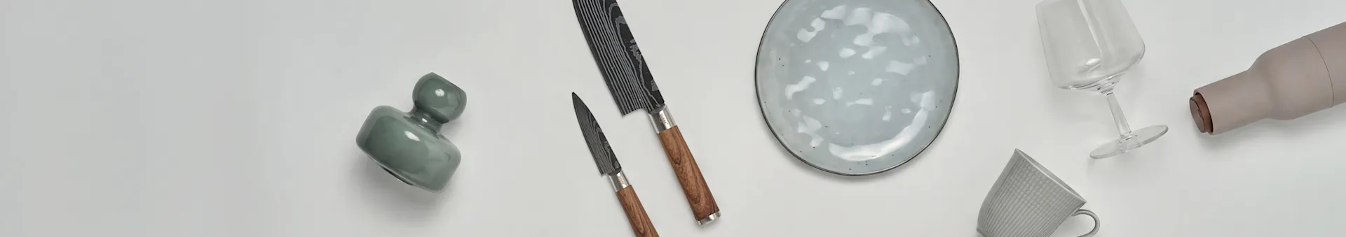 Vari articoli da cucina e da pranzo, inclusi un vaso verde, due coltelli, un piatto maculato, un calice da vino, una bottiglia beige e una tazza a motivi.