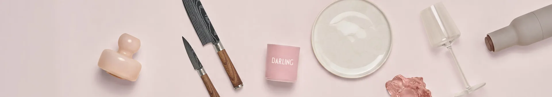 Assortimento di articoli da cucina e da pranzo su sfondo rosa: una scultura rosa, due coltelli decorati, un portacandele "DARLING", un piatto color crema, un bicchiere da vino trasparente e una bottiglia grigia.