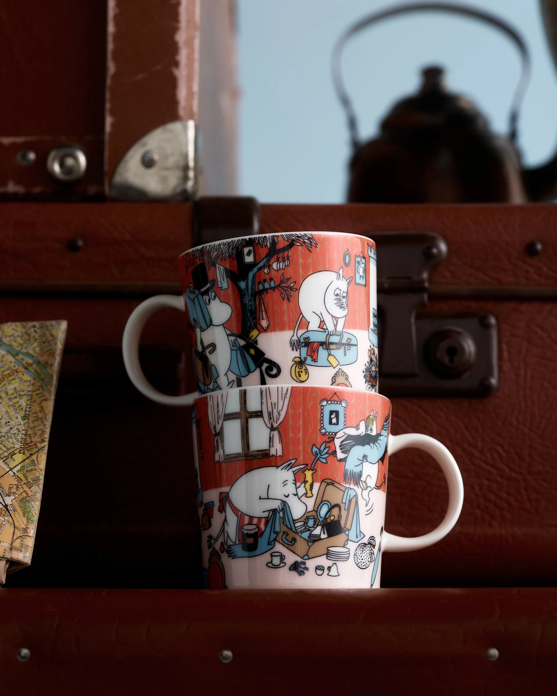 Due tazze Moomin con illustrazioni di personaggi rosso-arancio, impilate davanti a valigie marroni e una mappa sfocata.